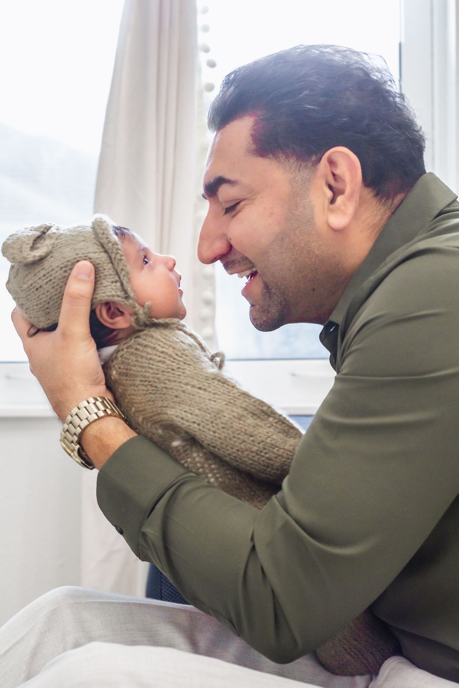 A smiling father holds his newborn baby wrapped in a knit swaddle and bear hat, sharing a tender moment indoors.