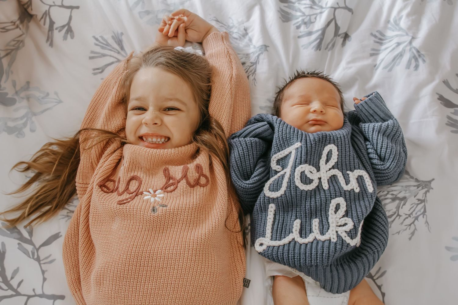 Two siblings, a smiling toddler girl and newborn baby boy named John Luk, wearing personalized knit sweaters on a bed.