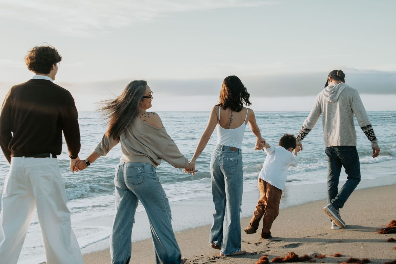 Family holding hands walking along a sandy beach near ocean waves at sunset in casual jeans and tops.