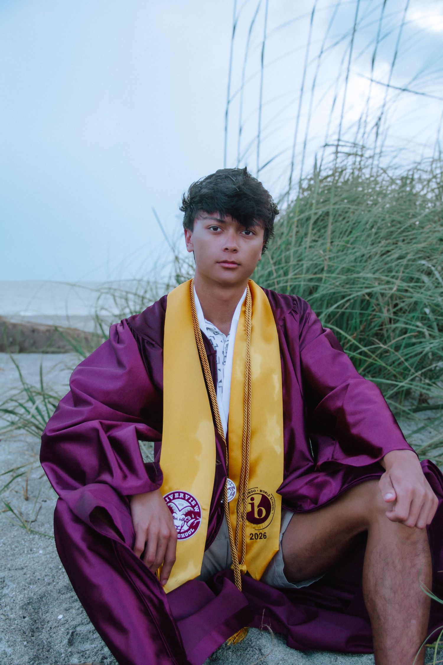 Young man in maroon graduation gown with gold stole sits on sandy beach with tall grass backdrop.