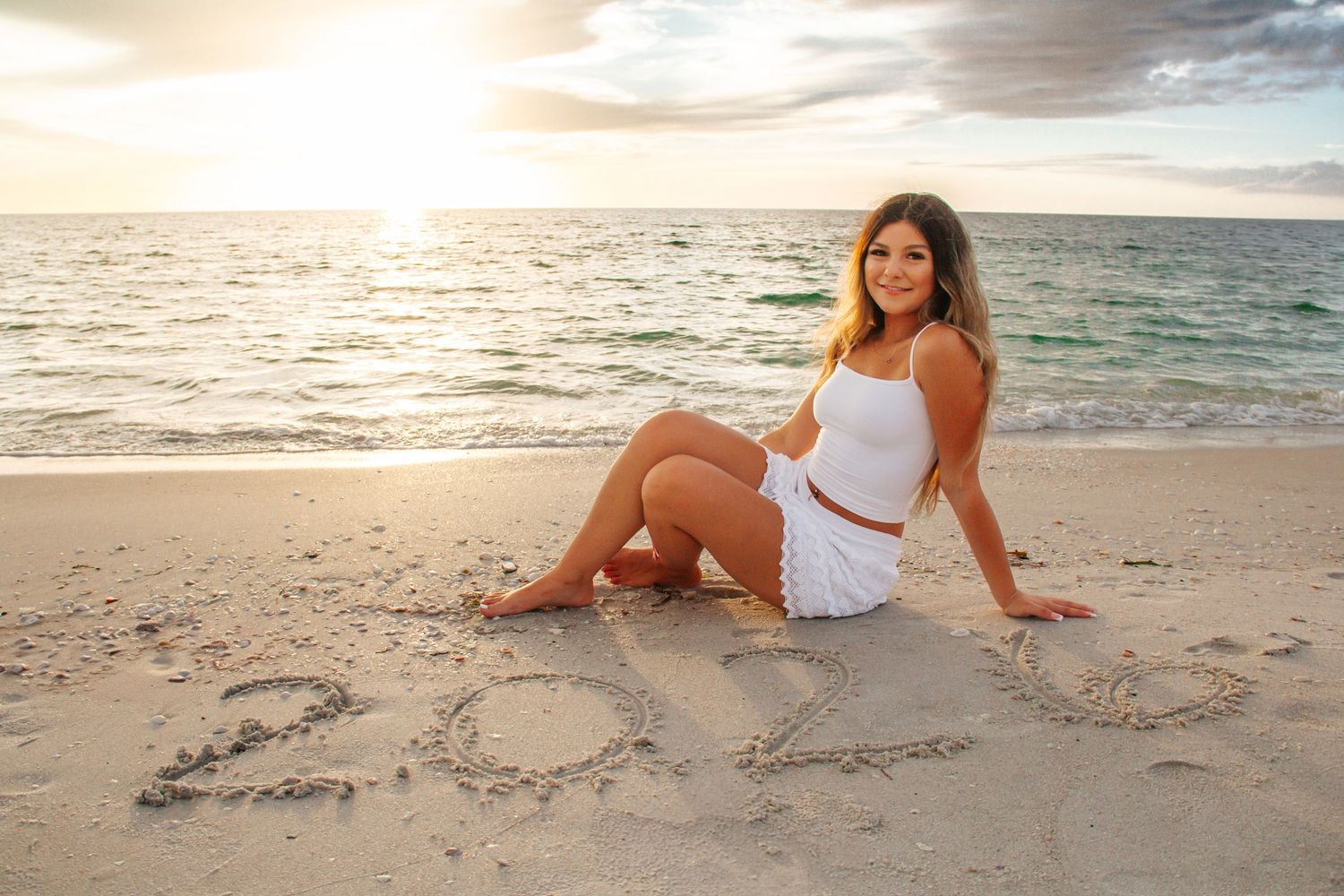 Young woman in white outfit sitting on sandy beach at sunset with 2023 written in the sand behind her.