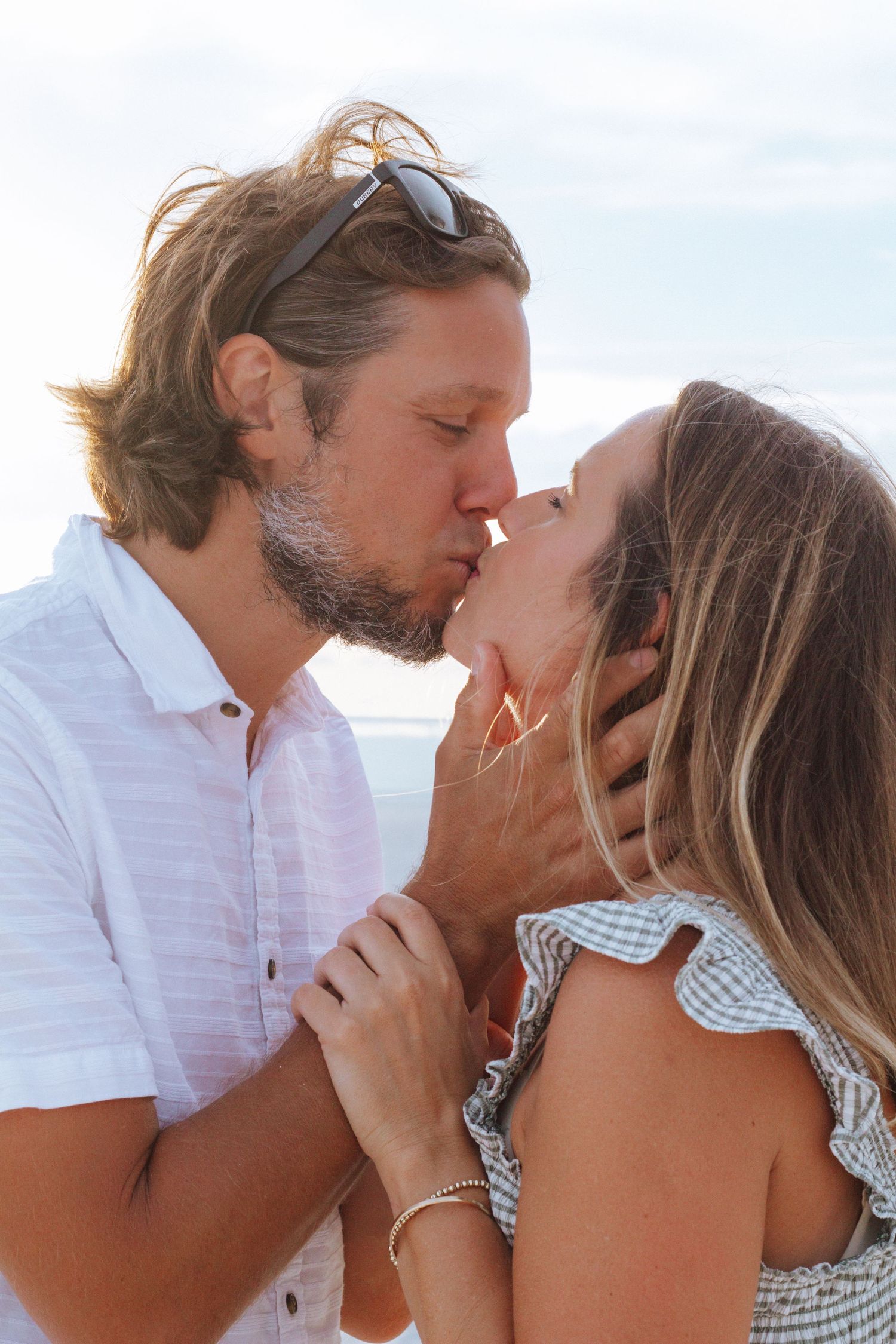 Romantic couple sharing a tender kiss on the beach at sunset, man in white shirt with sunglasses on head.