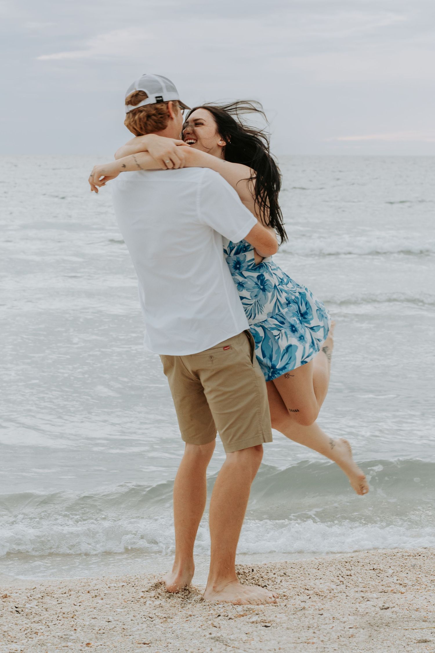 Couple embracing joyfully on a sandy beach, woman in floral dress lifted by man in white tee and khaki shorts.