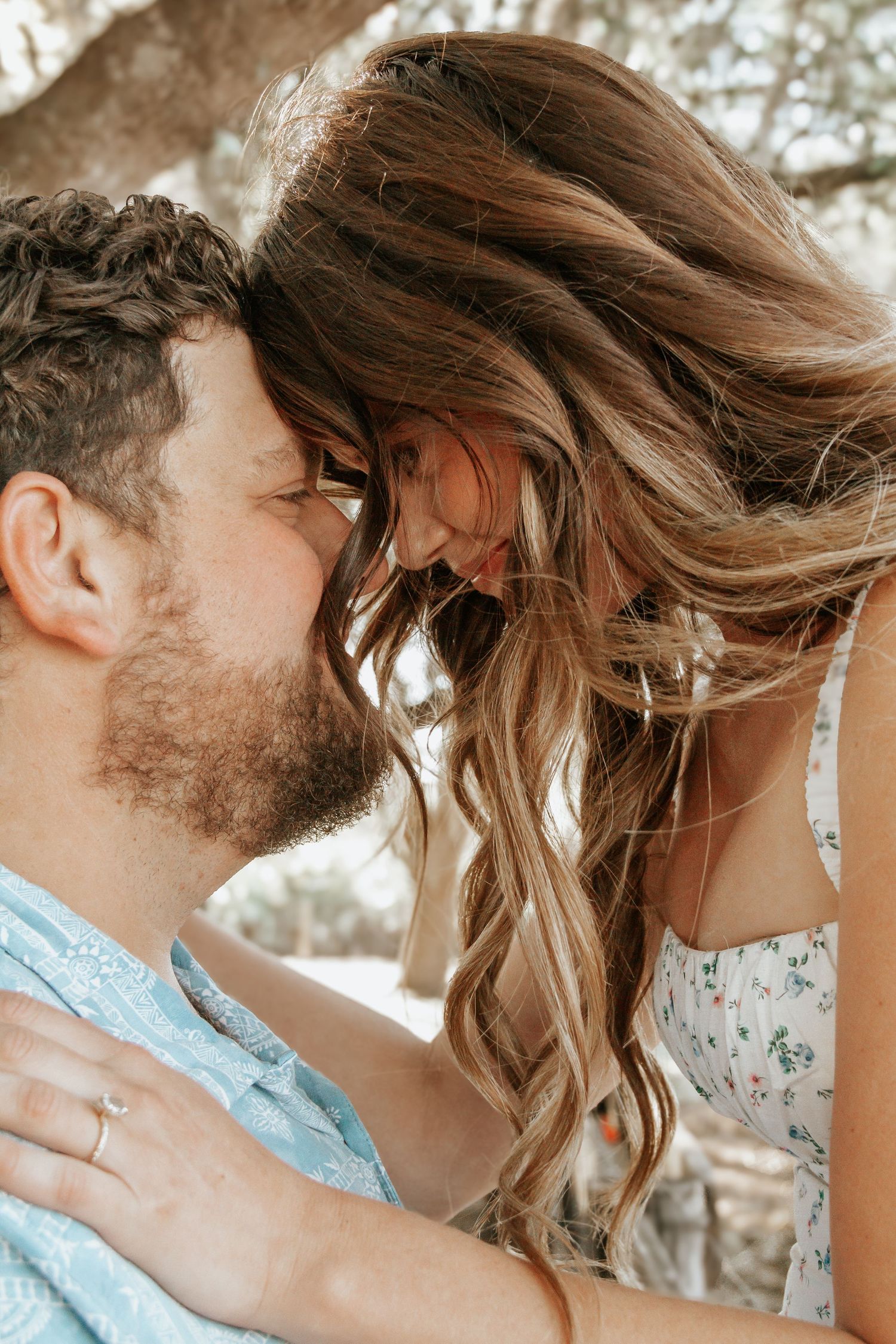 A couple sharing a romantic close moment, the woman leaning in toward the bearded man outdoors under a tree.