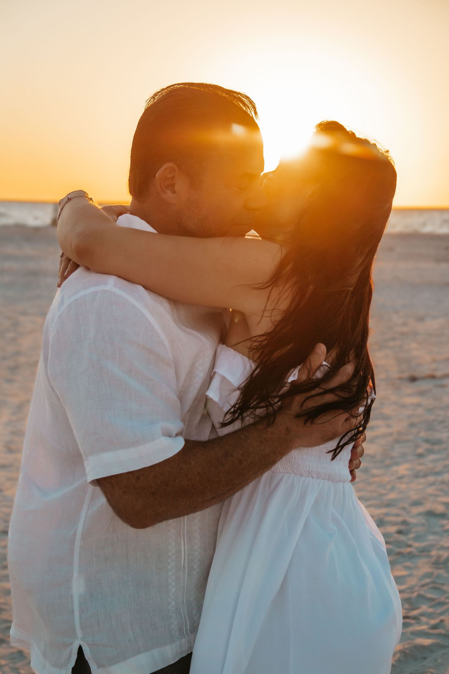 Couple sharing a romantic kiss on the beach at golden sunset, both dressed in white outfits.