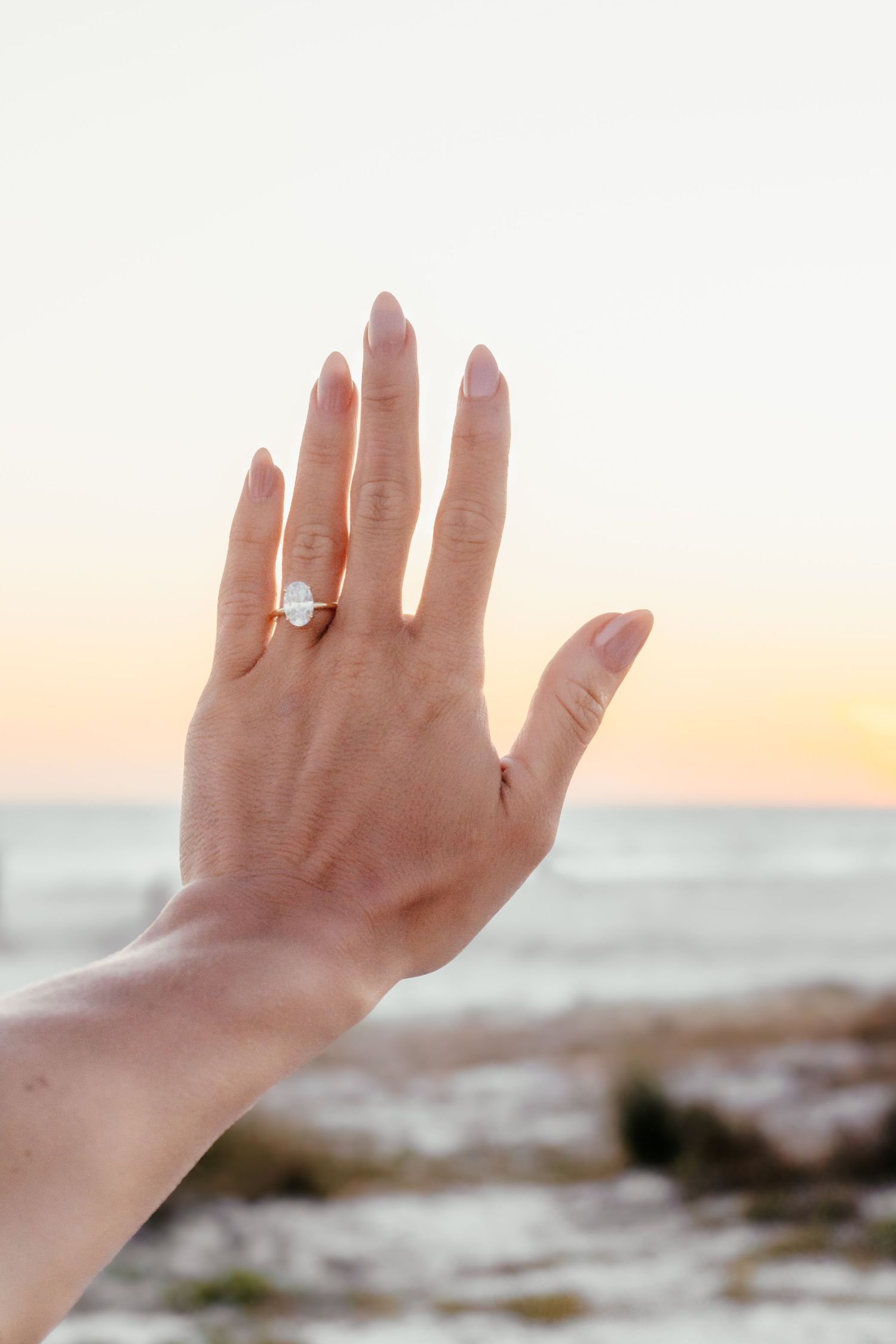 Hand raised against sunset ocean backdrop displaying elegant solitaire engagement ring with clear stone on ring finger.