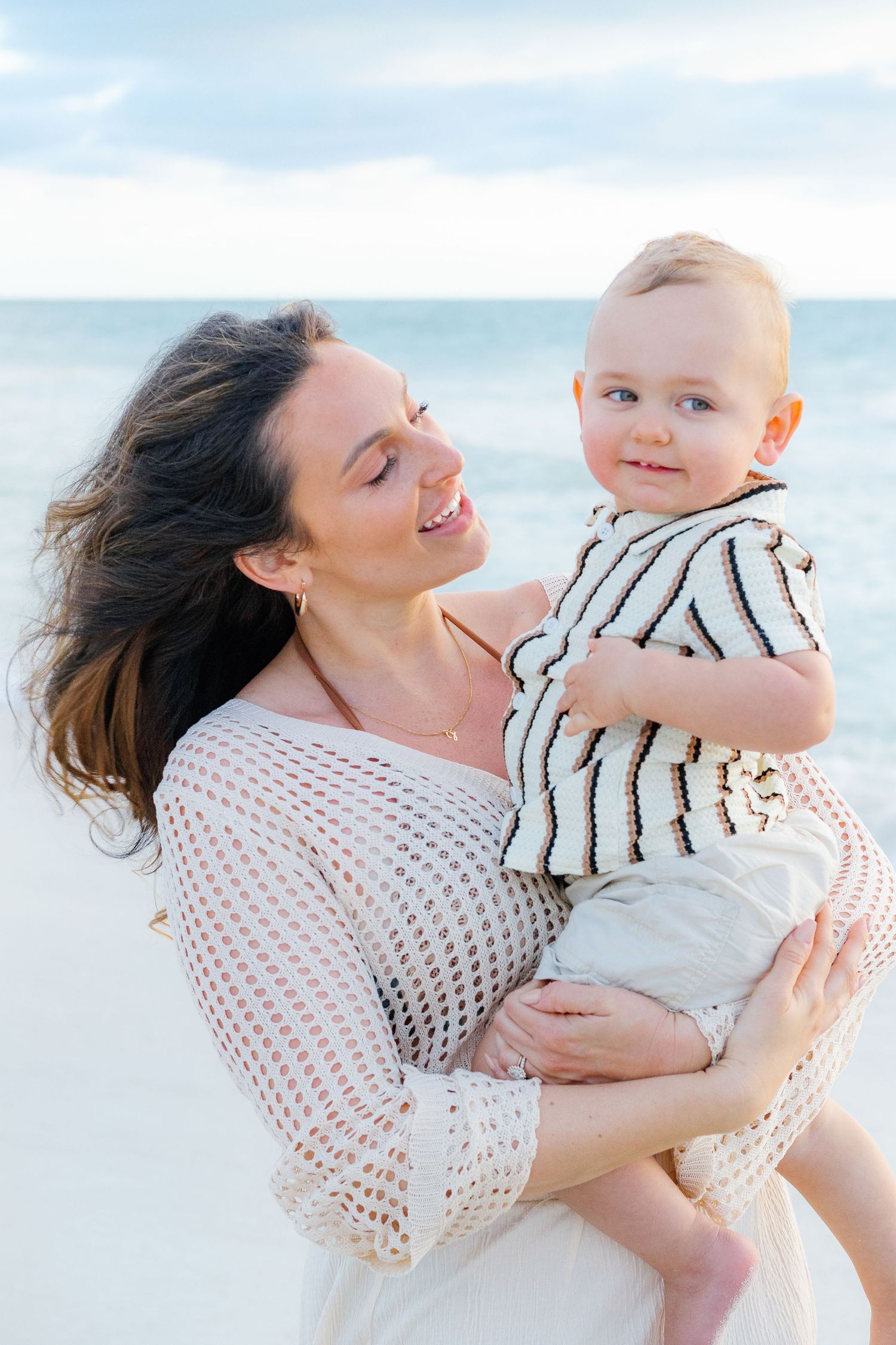 A smiling mother holds her happy baby on a sunny beach with ocean waves in the background.