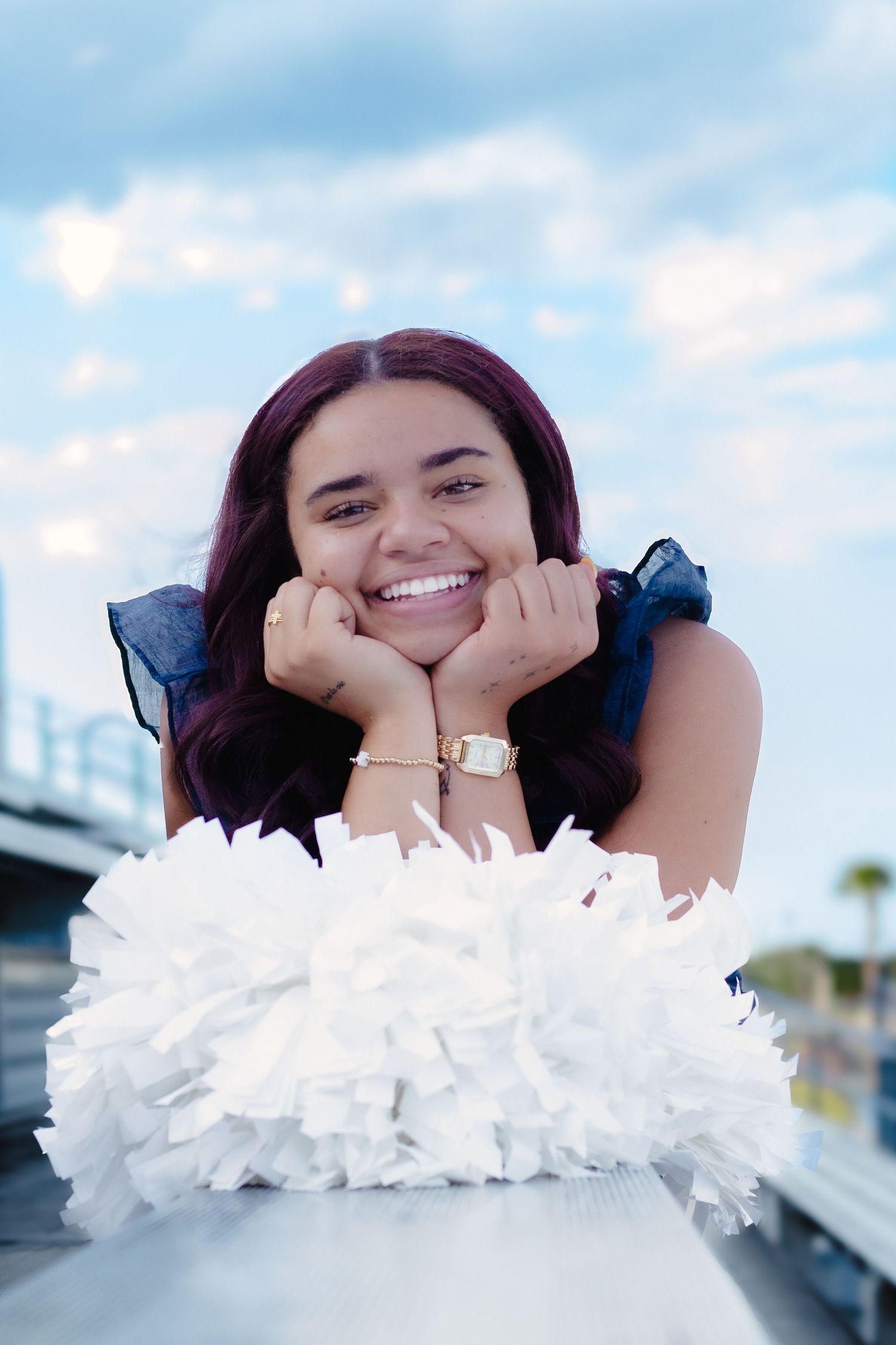 Happy young woman with purple hair leaning on a railing, smiling brightly outdoors under a cloudy sky.