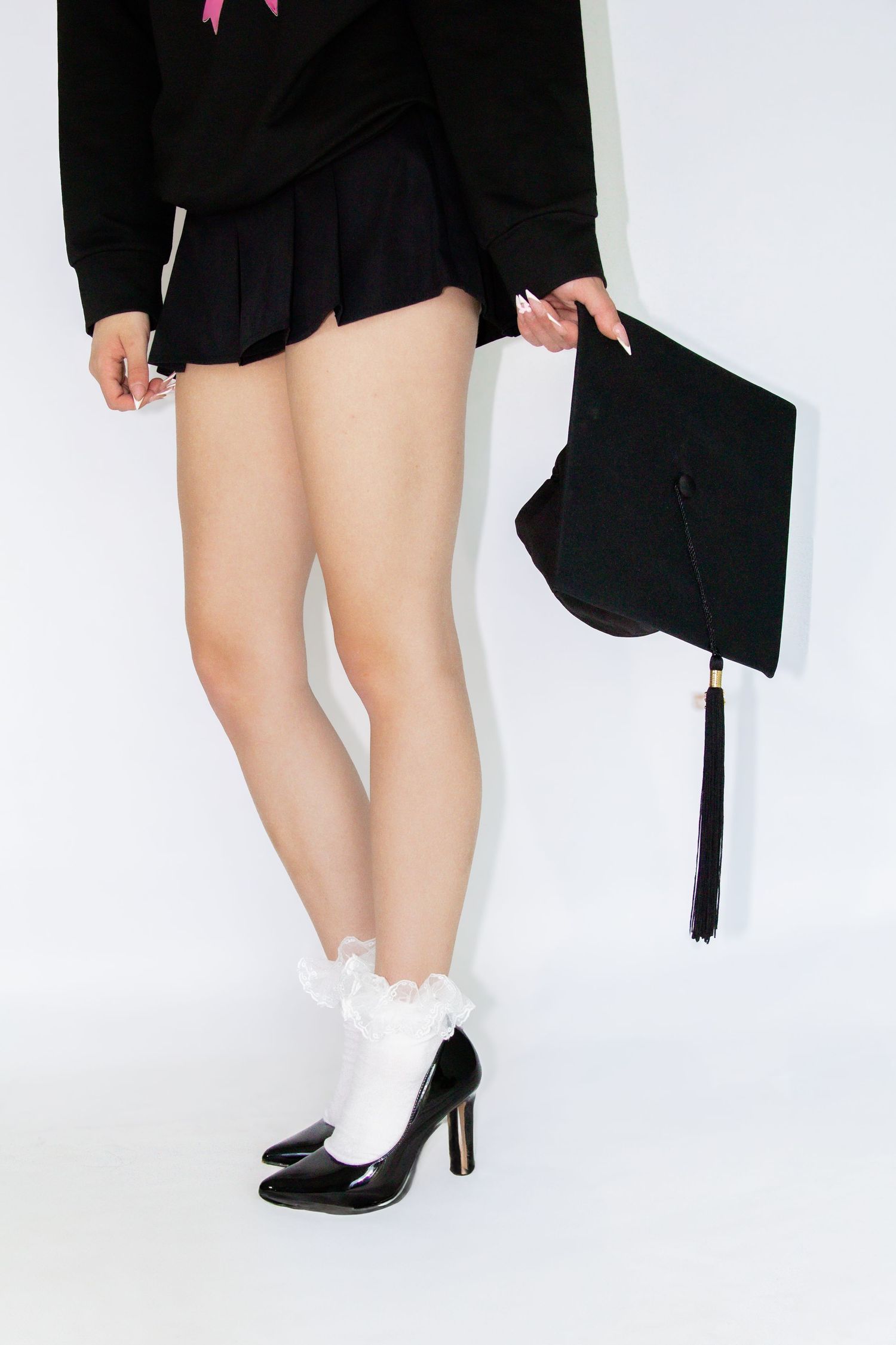Woman in black mini skirt and heels holding a graduation cap, wearing white frilly socks against white background.