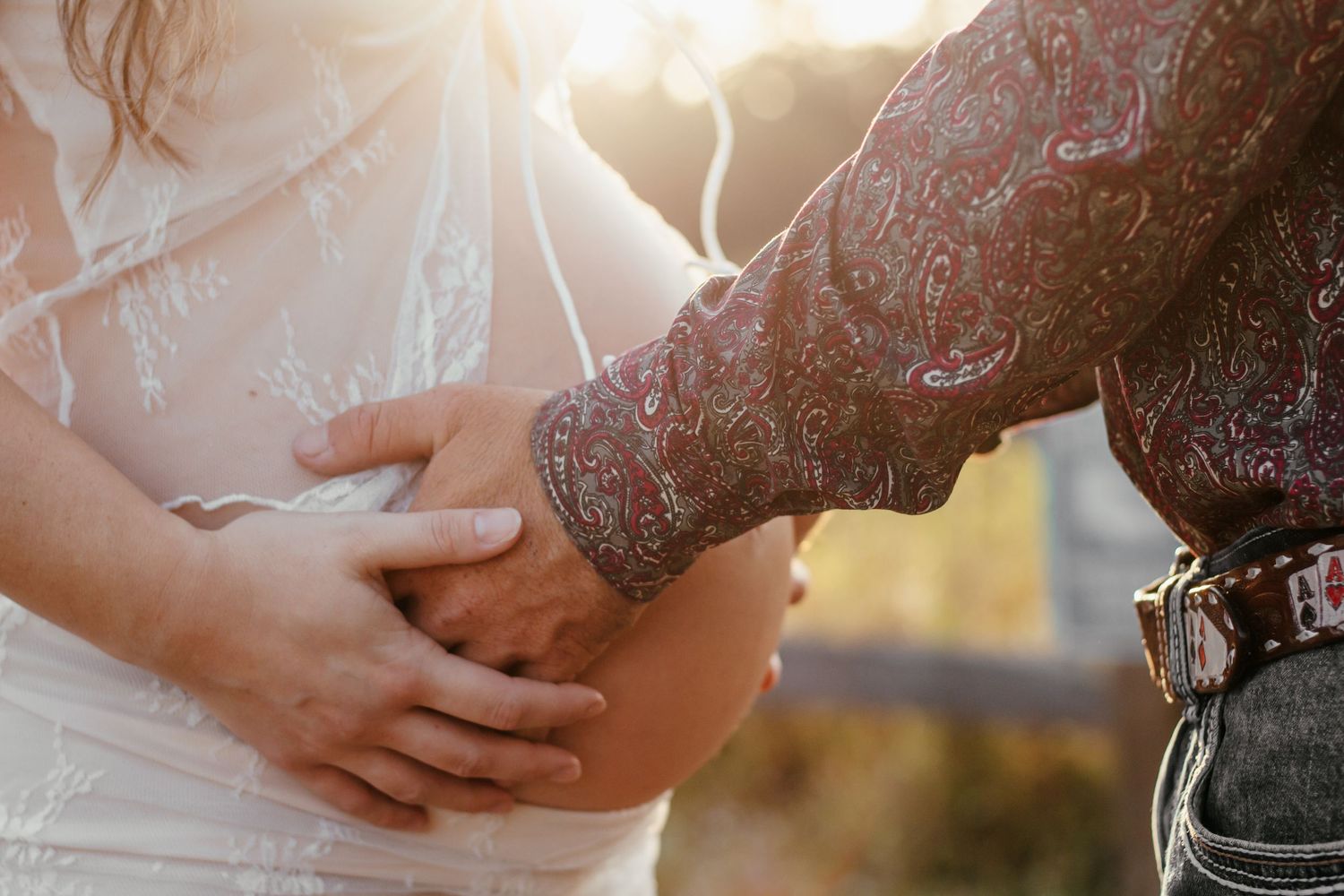 Couple tenderly cradling pregnant belly in golden sunlight, woman in lace dress, man in paisley shirt with leather belt.