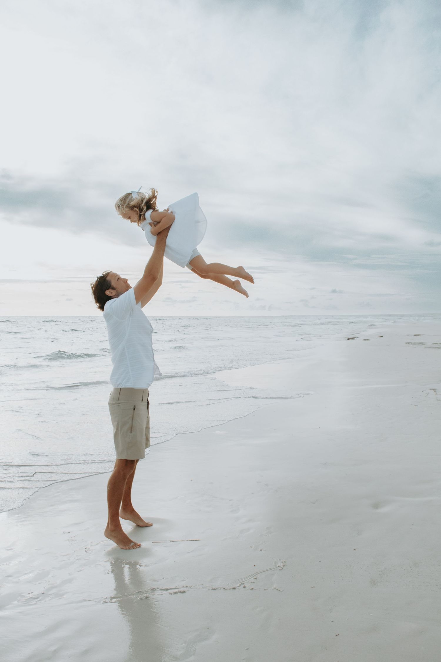 Father joyfully lifting child high in the air on a serene beach with gentle waves and cloudy sky.