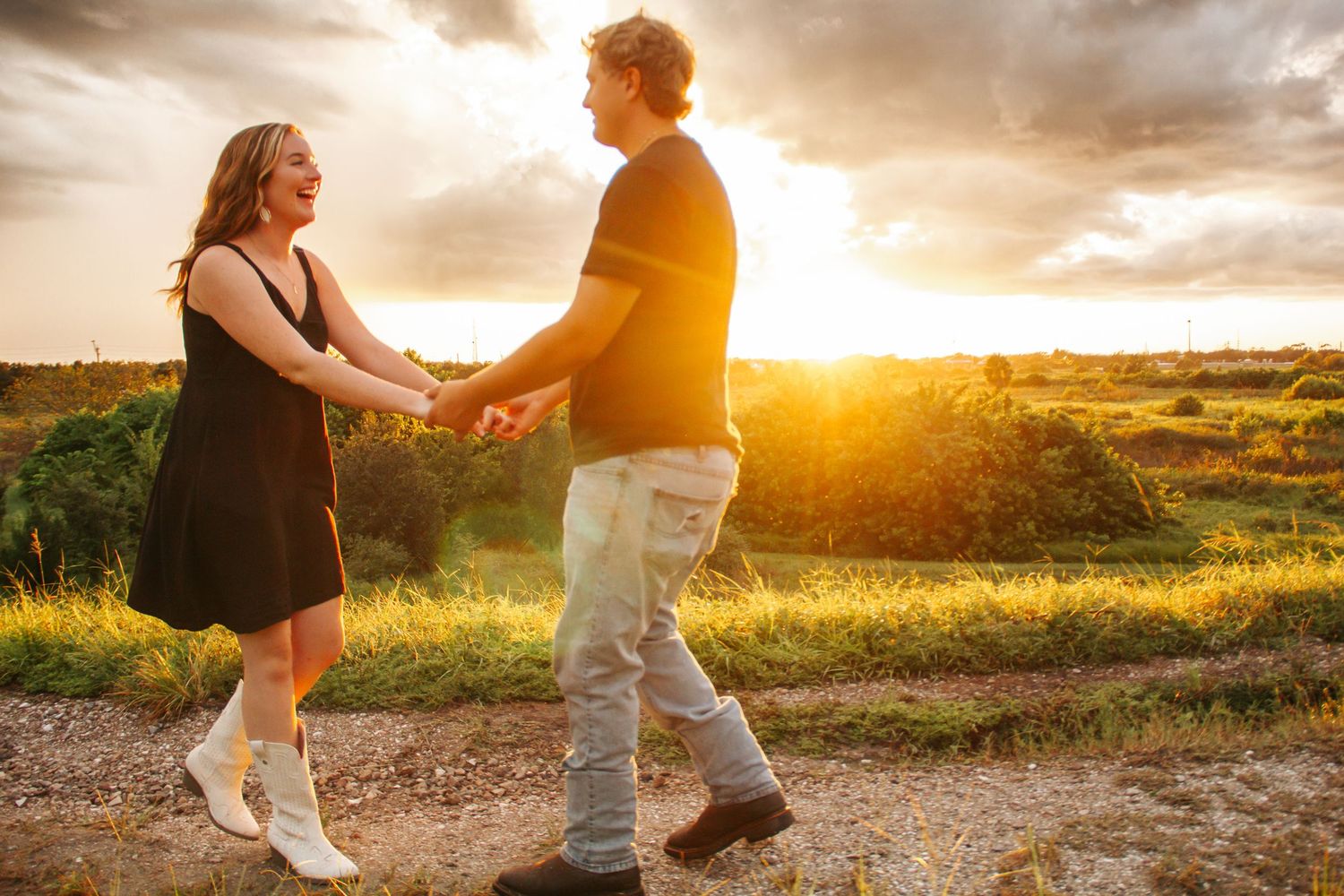 Couple holding hands and dancing at golden sunset on a scenic dirt path surrounded by lush greenery.