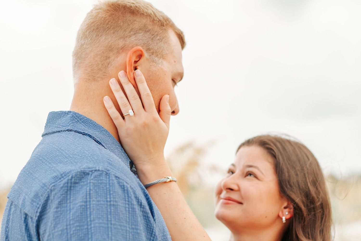 A smiling woman tenderly touches a man's face outdoors, showcasing an engagement ring on her finger.