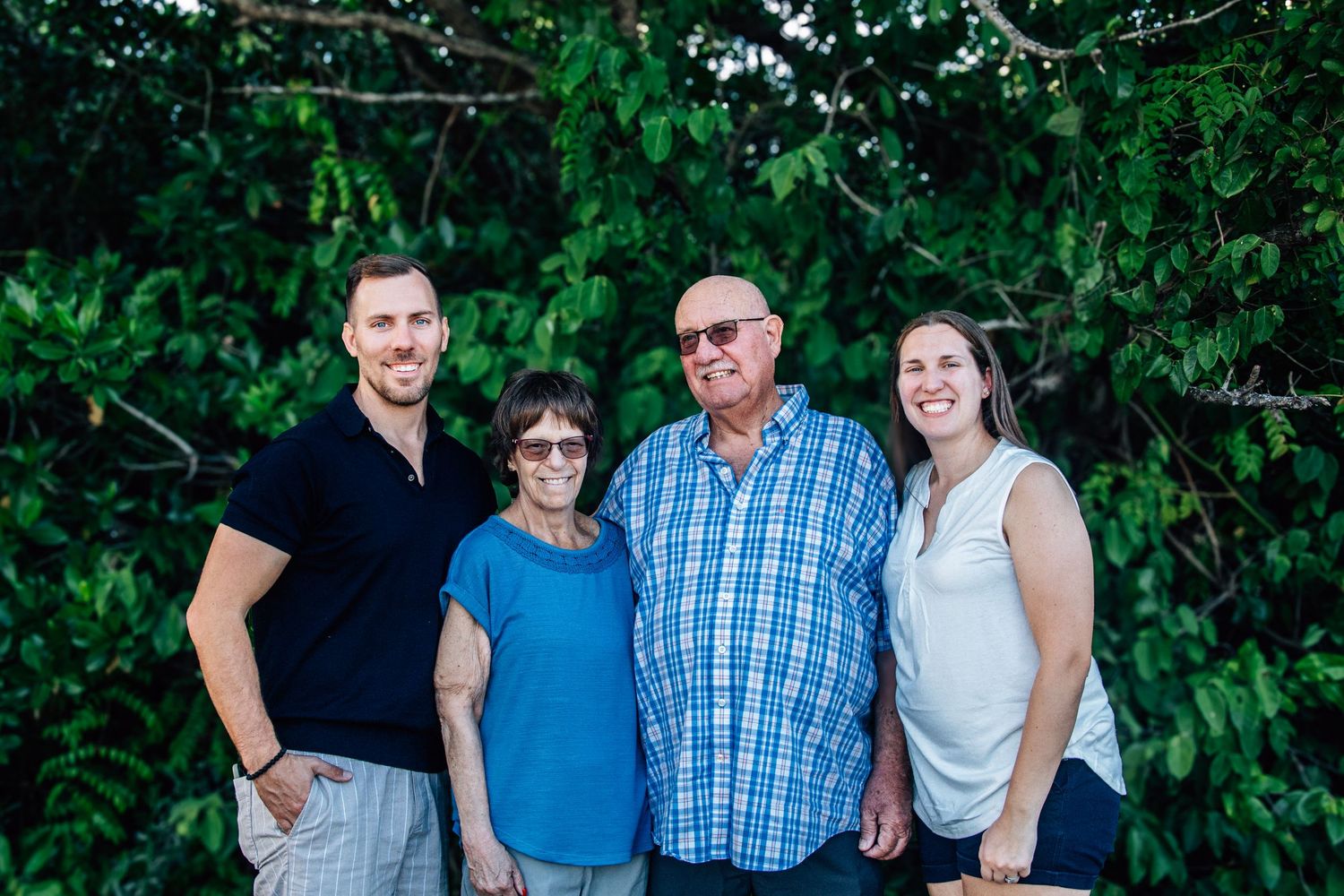 Family of four smiling outdoors with lush green trees in the background on a sunny day.