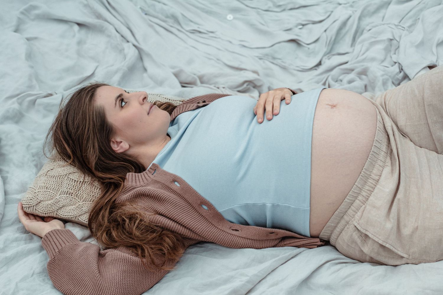 Pregnant woman lying on bed in blue top, resting with hand on baby bump, looking upward peacefully.