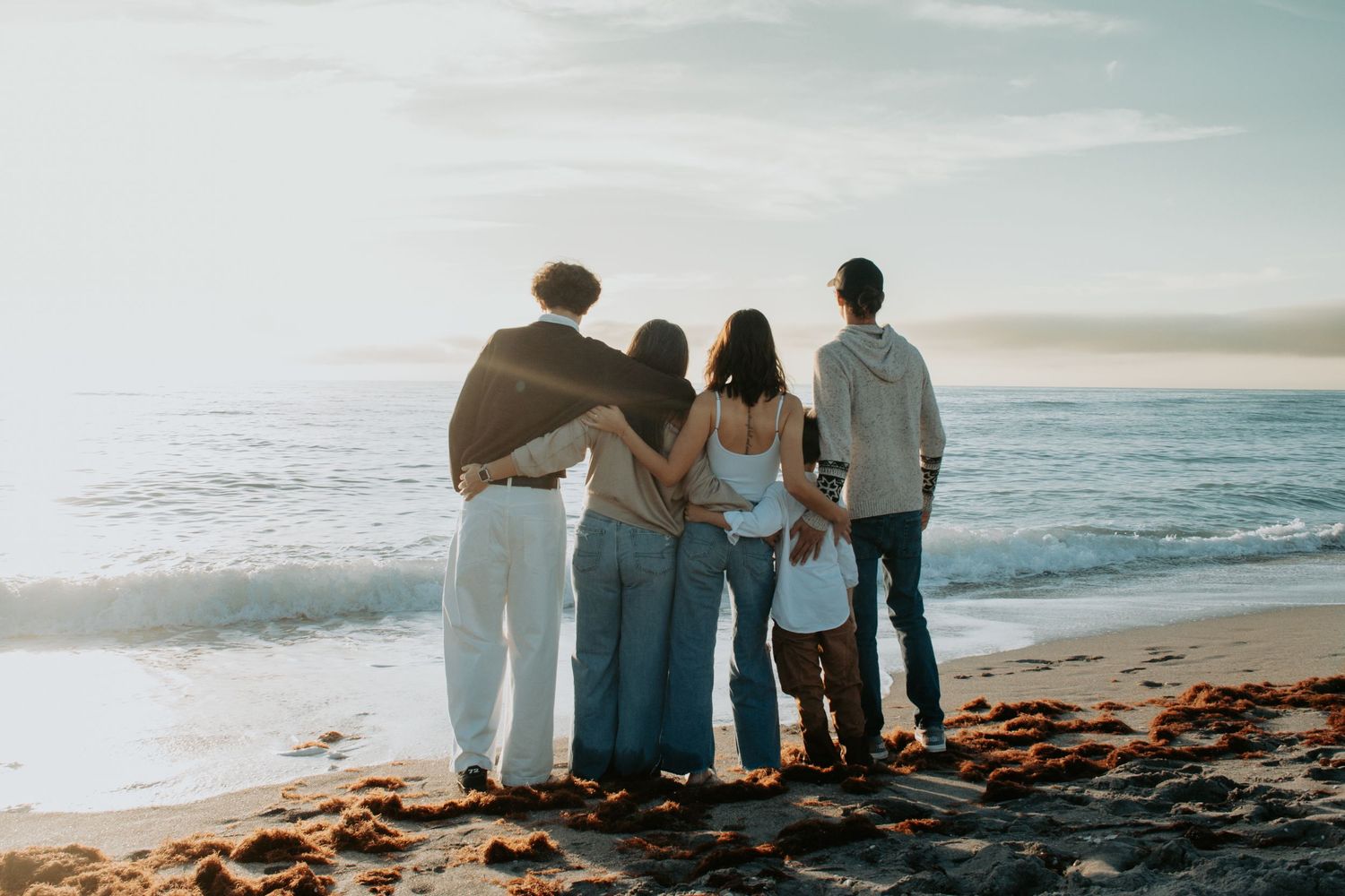 Family of five standing together on a sandy beach, viewed from behind, watching ocean waves at sunset.
