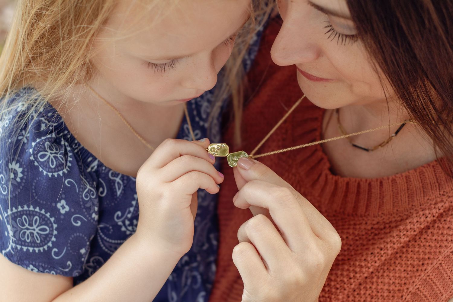 A mother and young daughter closely examine a delicate gold locket necklace together.