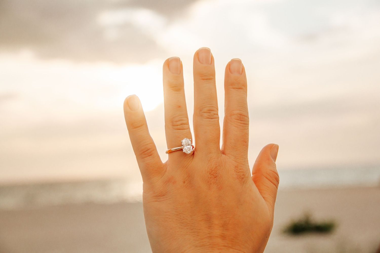 Hand raised against sunset beach backdrop displaying elegant solitaire diamond engagement ring on ring finger.