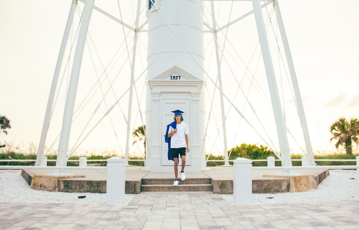 Graduate in cap and gown poses proudly in front of a white lighthouse structure on a sunny day.