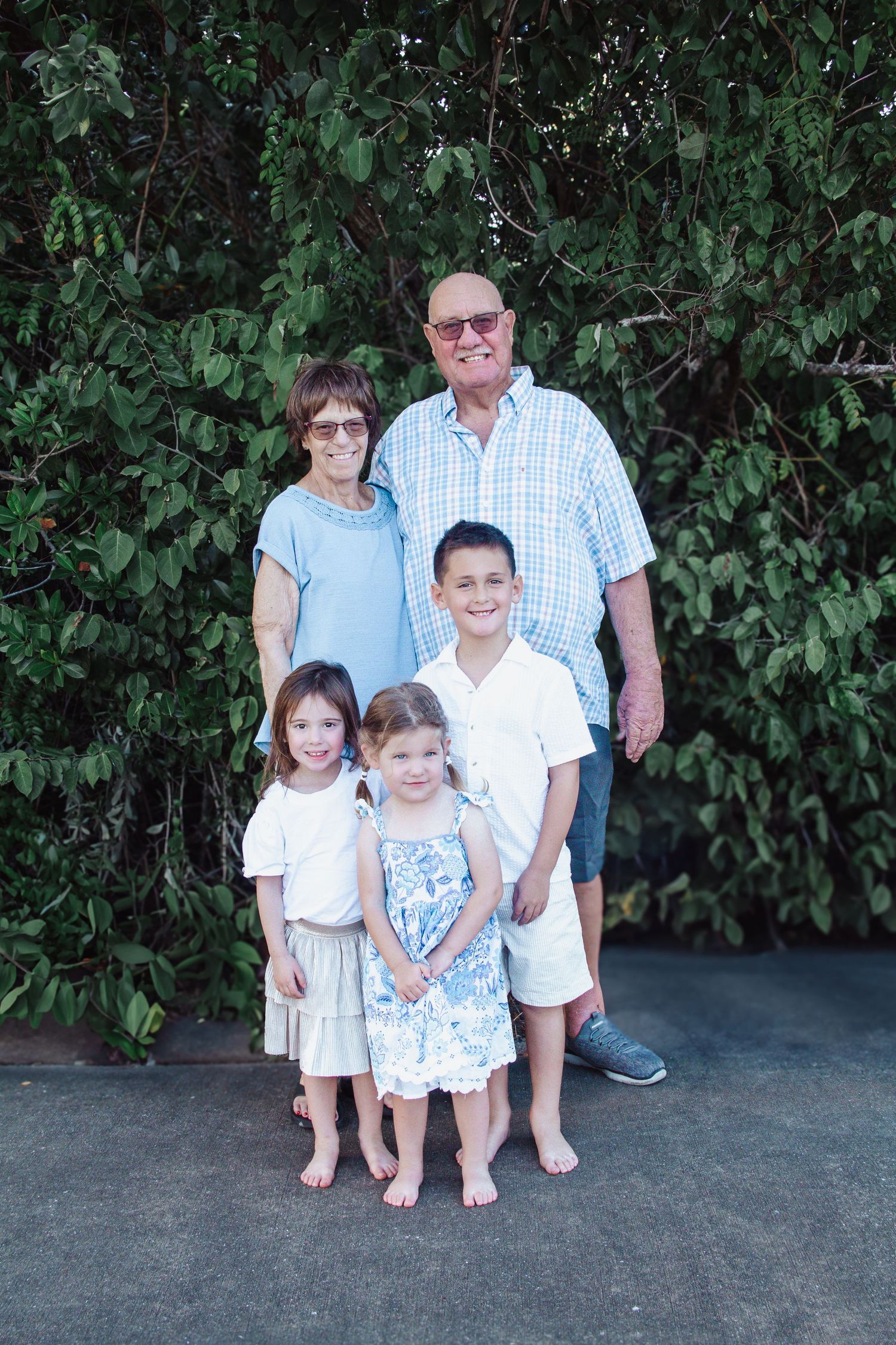 Grandparents posing outdoors with three young grandchildren in front of lush green trees.