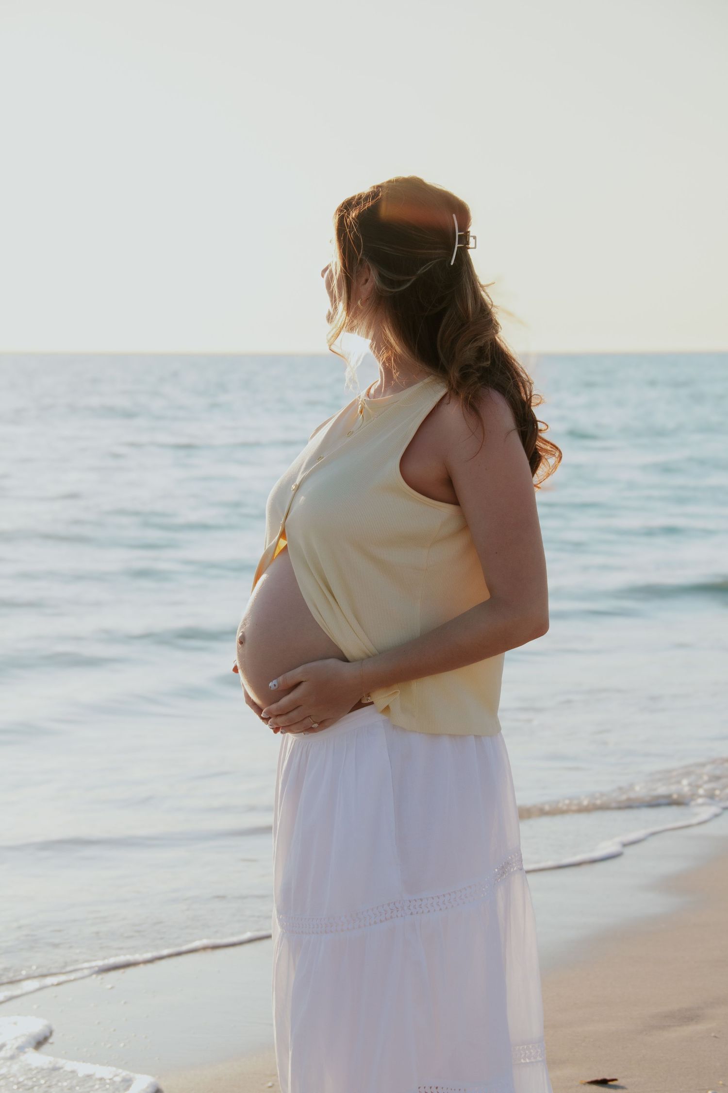 Pregnant woman in yellow top and white skirt cradling her baby bump while standing on a serene beach at sunset.