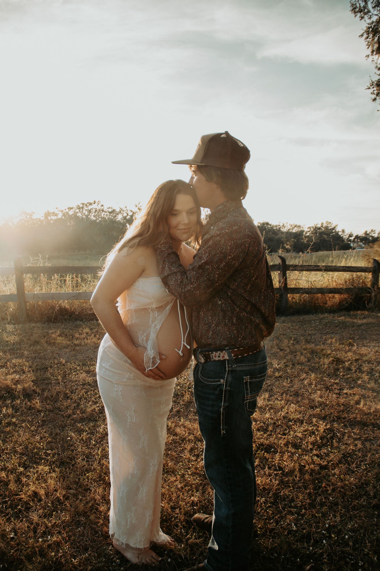 Couple sharing a tender moment at golden hour in a field, woman in white lace maternity dress, man in cowboy hat.