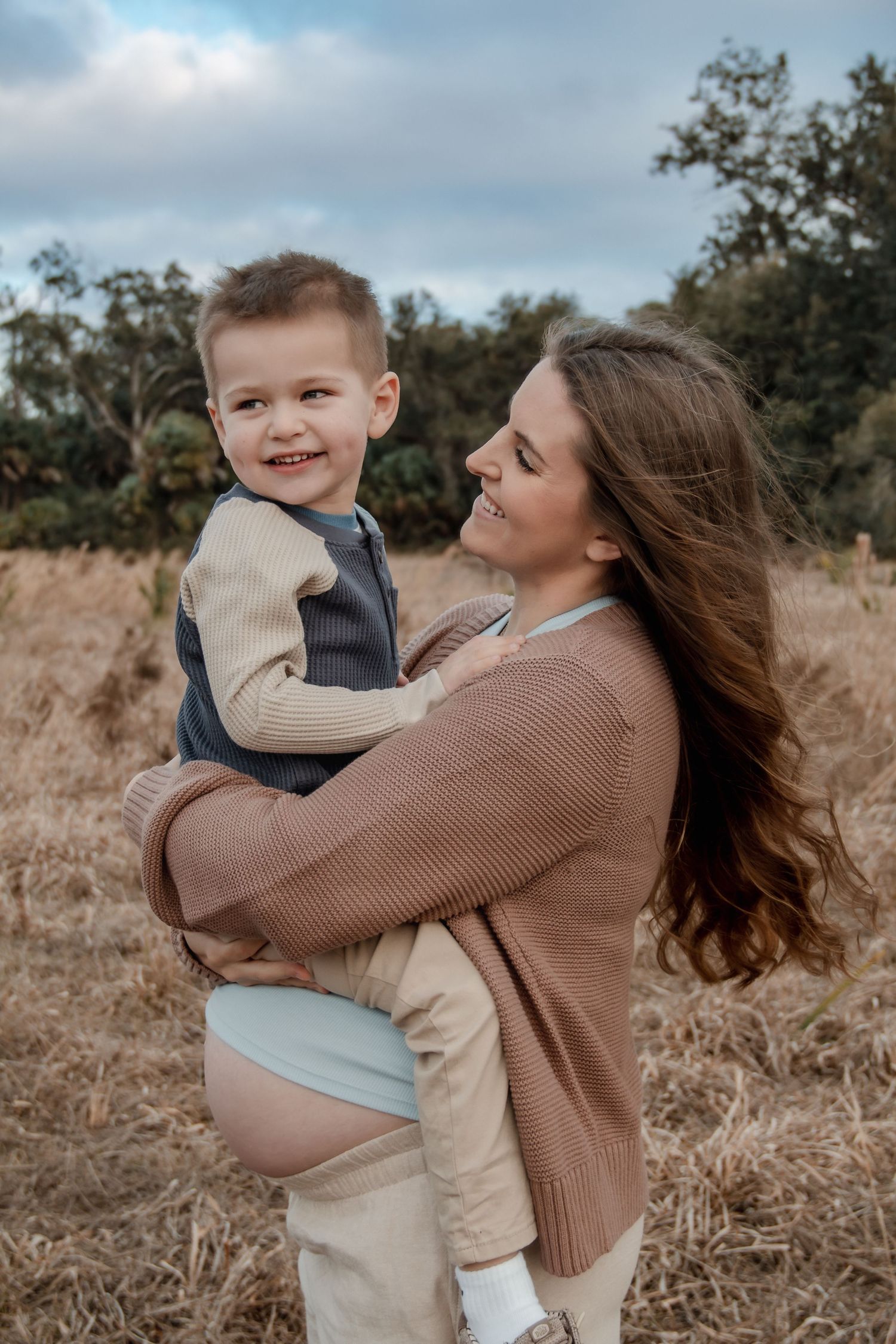 A smiling young boy being held by a woman in a brown sweater in an autumn field.
