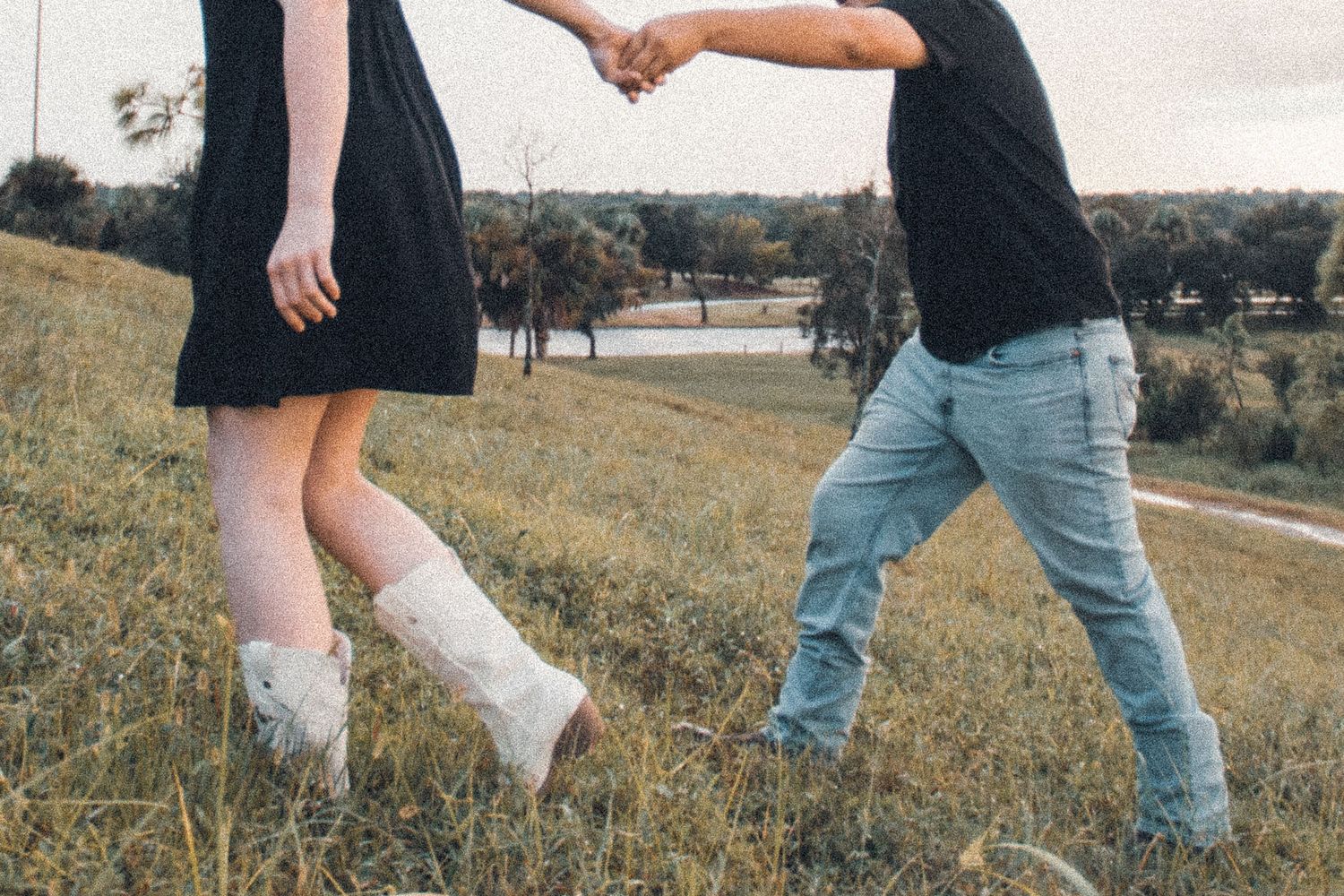 Couple holding hands in a grassy field, woman in black dress with white boots, man in jeans and dark shirt.