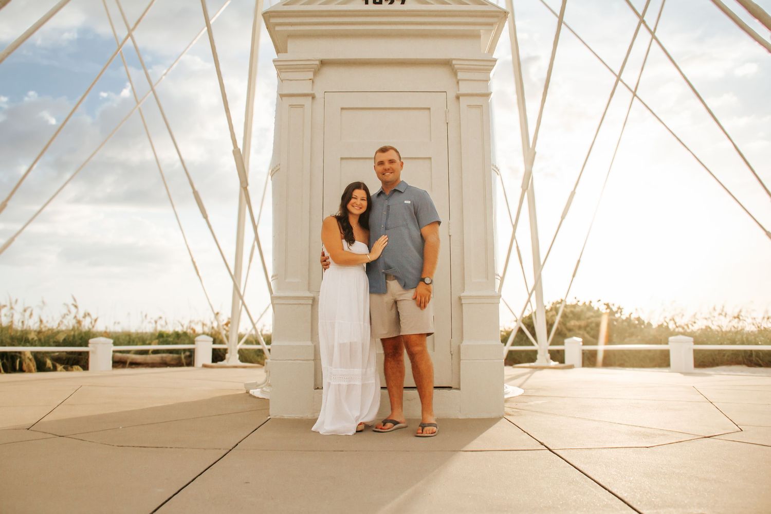 Couple posing together on a bridge at sunset, woman in white dress and man in blue shirt, with cable supports behind them.