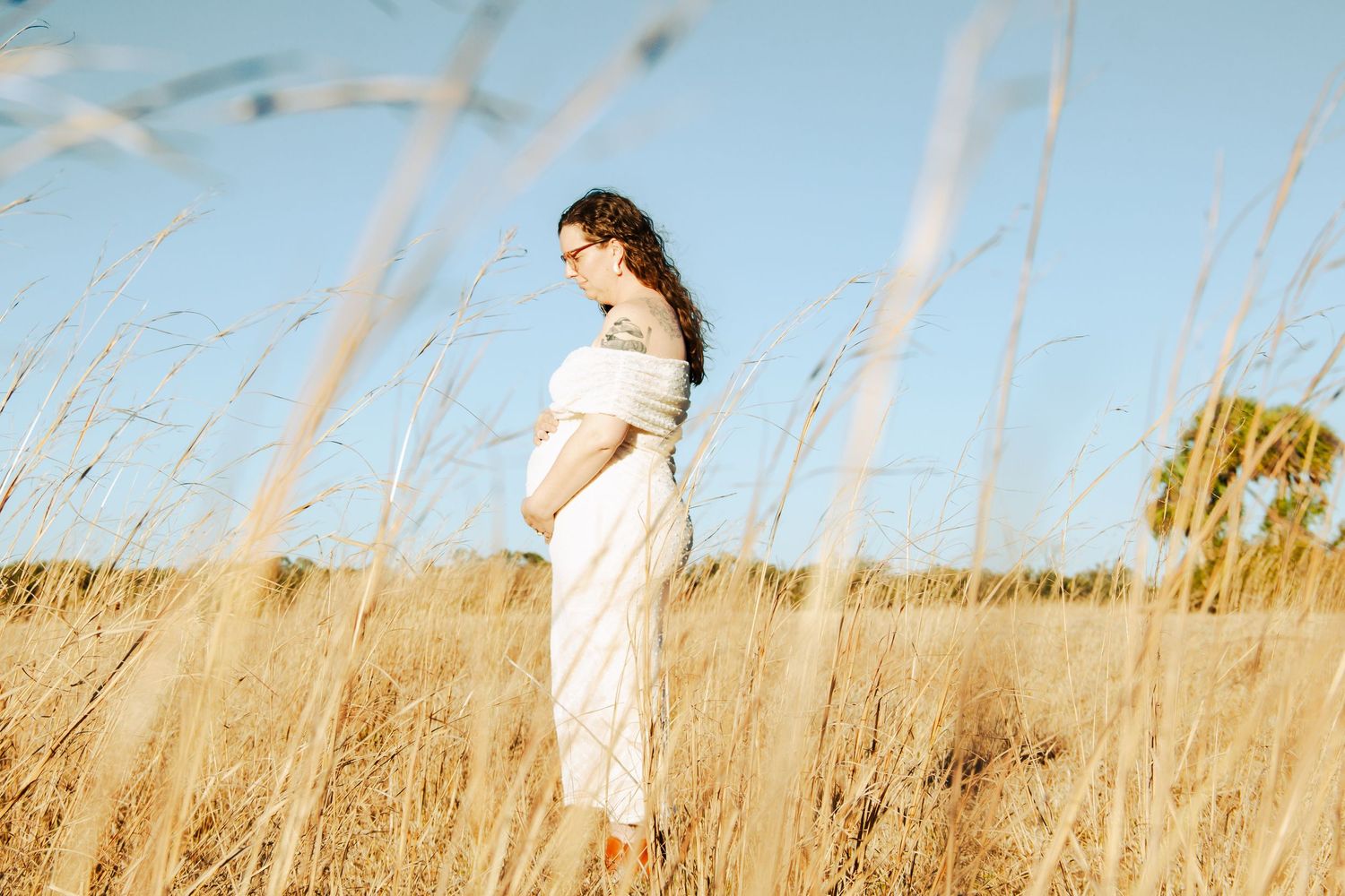 Pregnant woman in white dress standing in golden field under blue sky, cradling baby bump during outdoor maternity photoshoot.