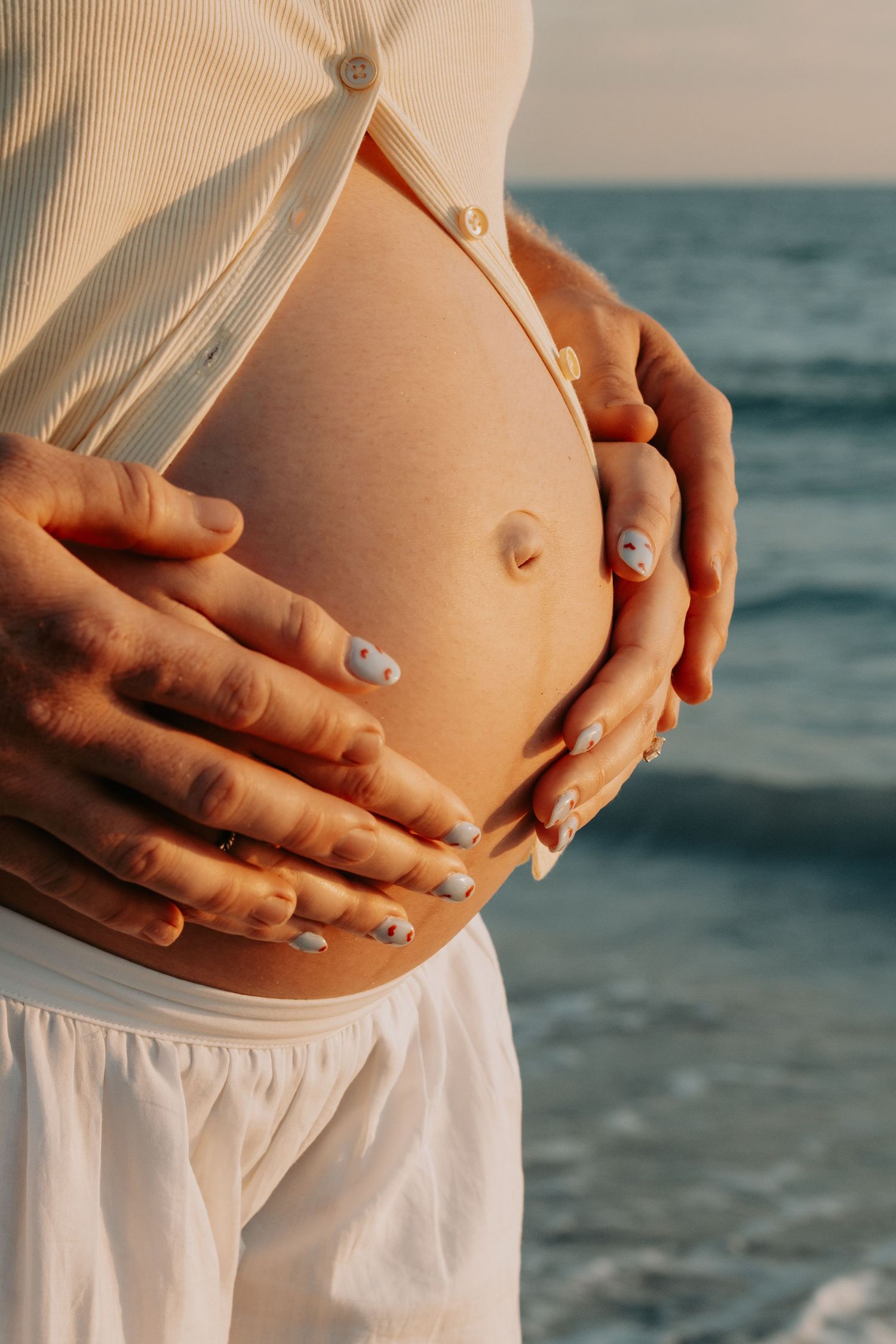 Pregnant woman's bare belly held by two pairs of hands near the ocean at sunset.