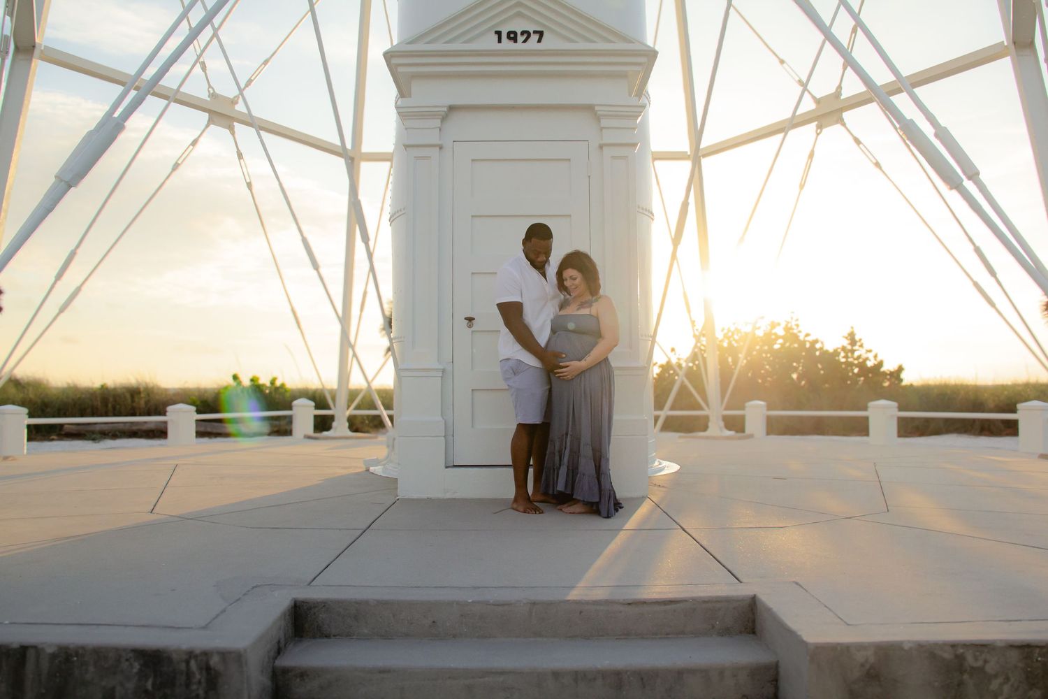 Couple poses at sunset by a white lighthouse door during a maternity photo session outdoors.