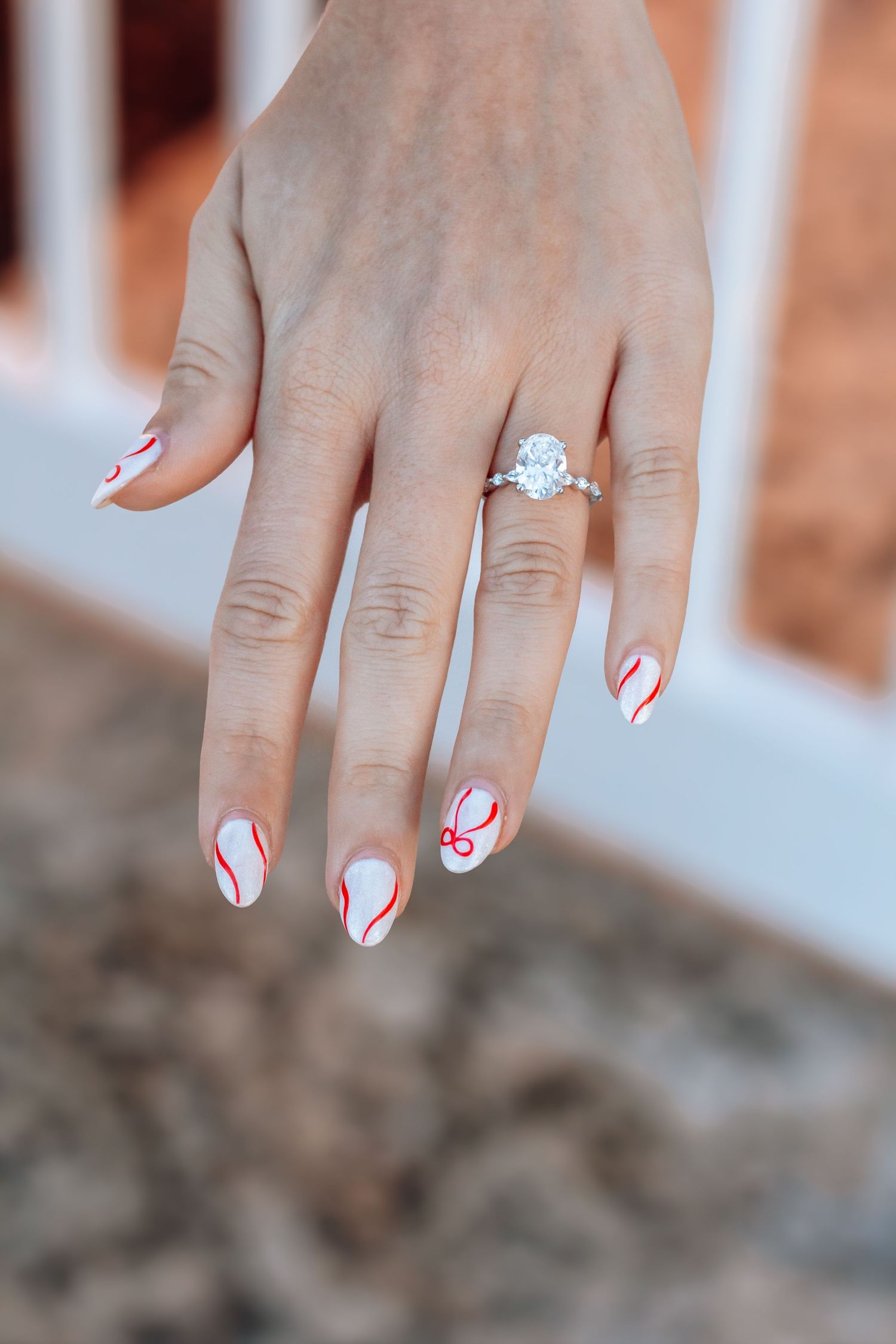 Close-up of a hand with white nails featuring red swirl nail art, wearing a stunning oval diamond engagement ring.