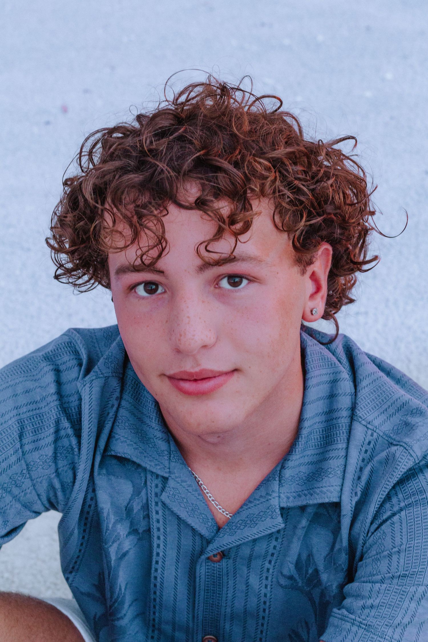 Young man with curly auburn hair, hoop earring, and blue shirt poses outdoors in a casual portrait photo.