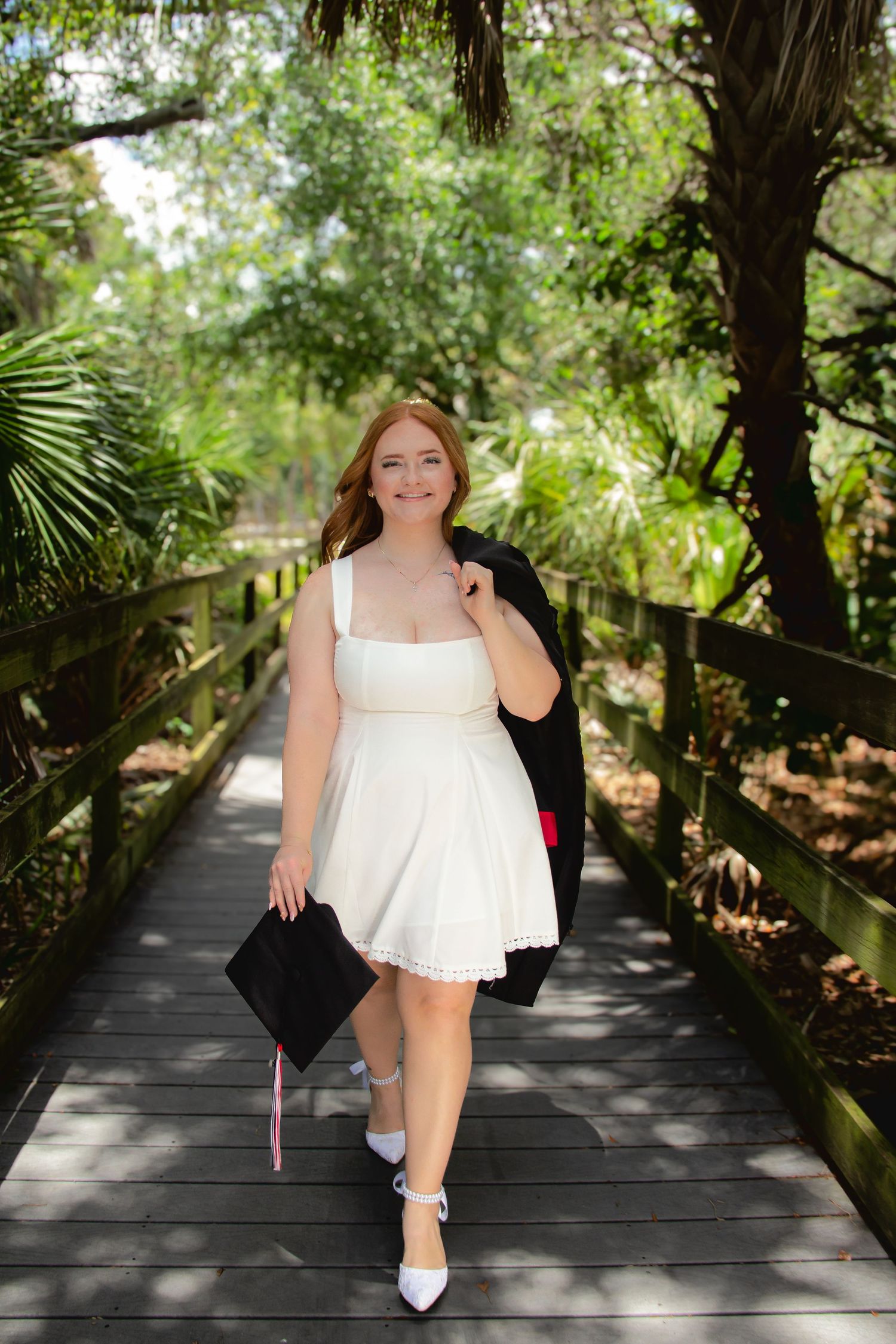 Young woman in white dress carrying graduation cap and gown walks along a lush green wooden boardwalk path.