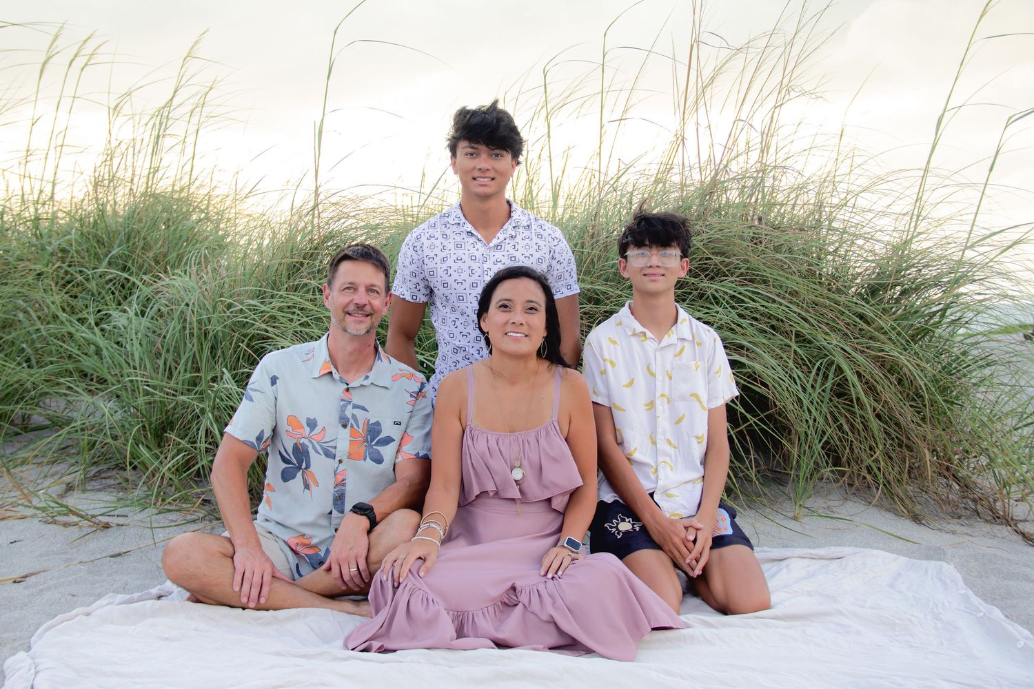 Family of four smiling together on a sandy beach with green dune grass backdrop.