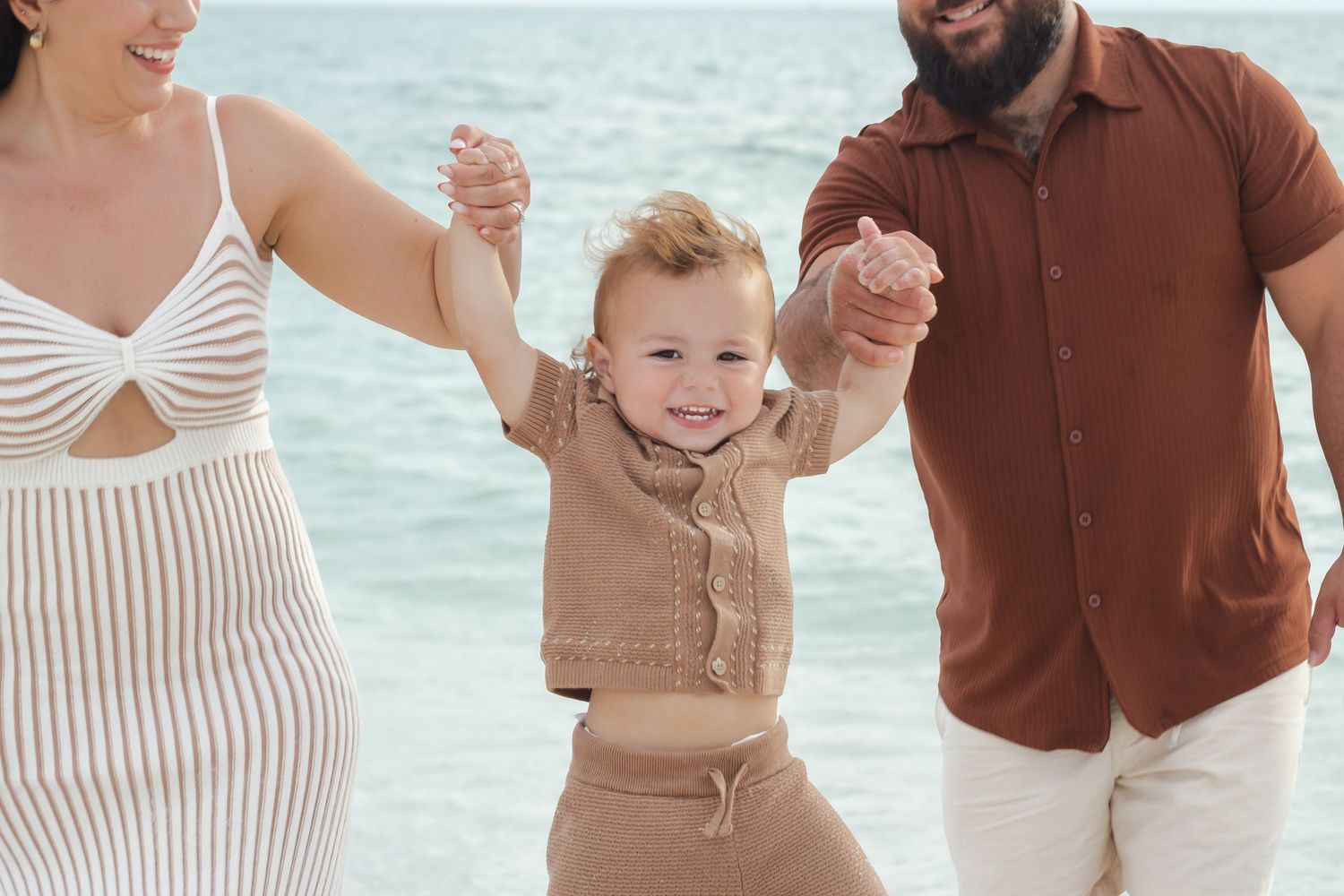 Happy toddler held by parents hands walking on a sunny beach with ocean waves in the background.