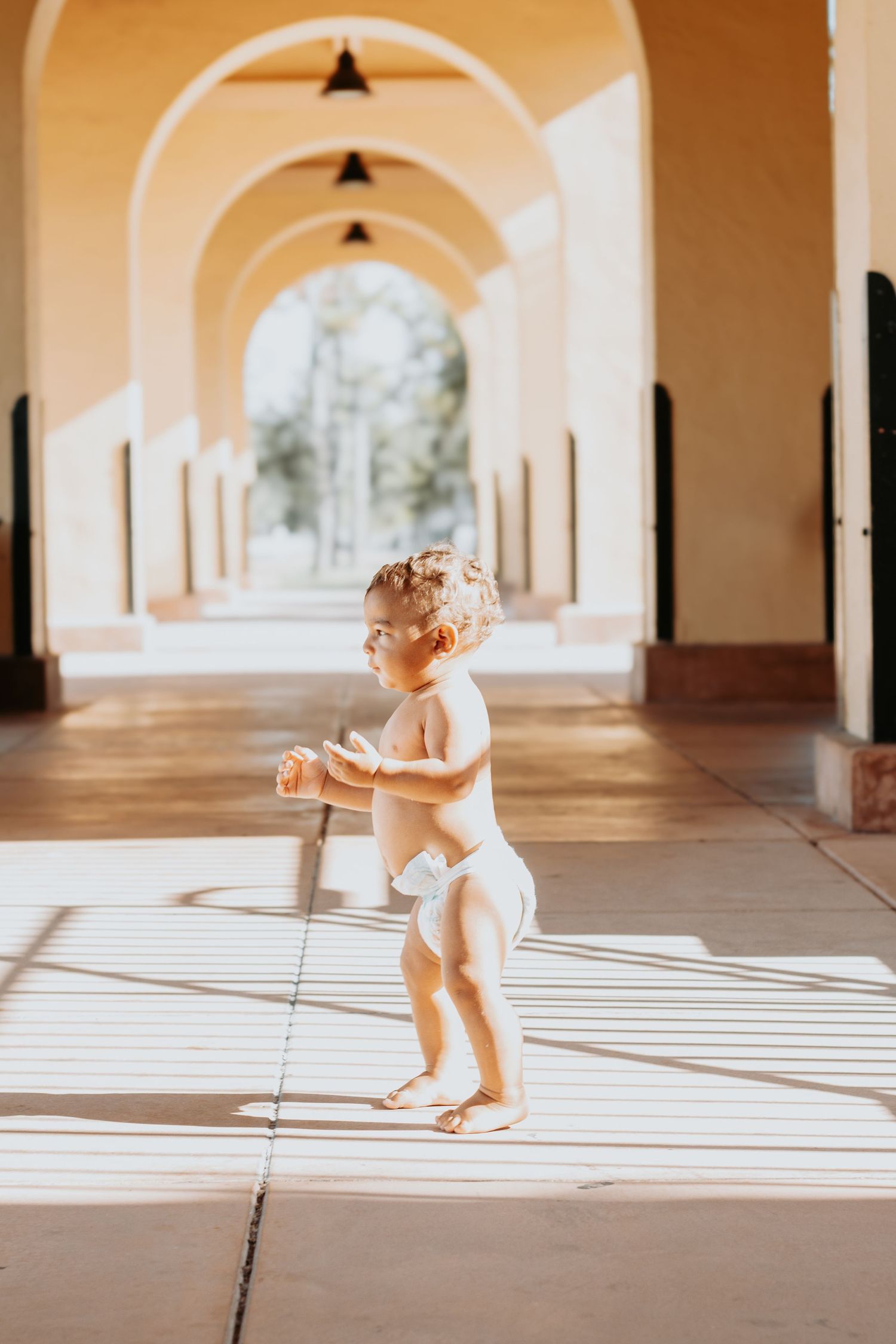 A small child walks through an elegant arched corridor casting long shadows in warm sunlight streaming through windows.