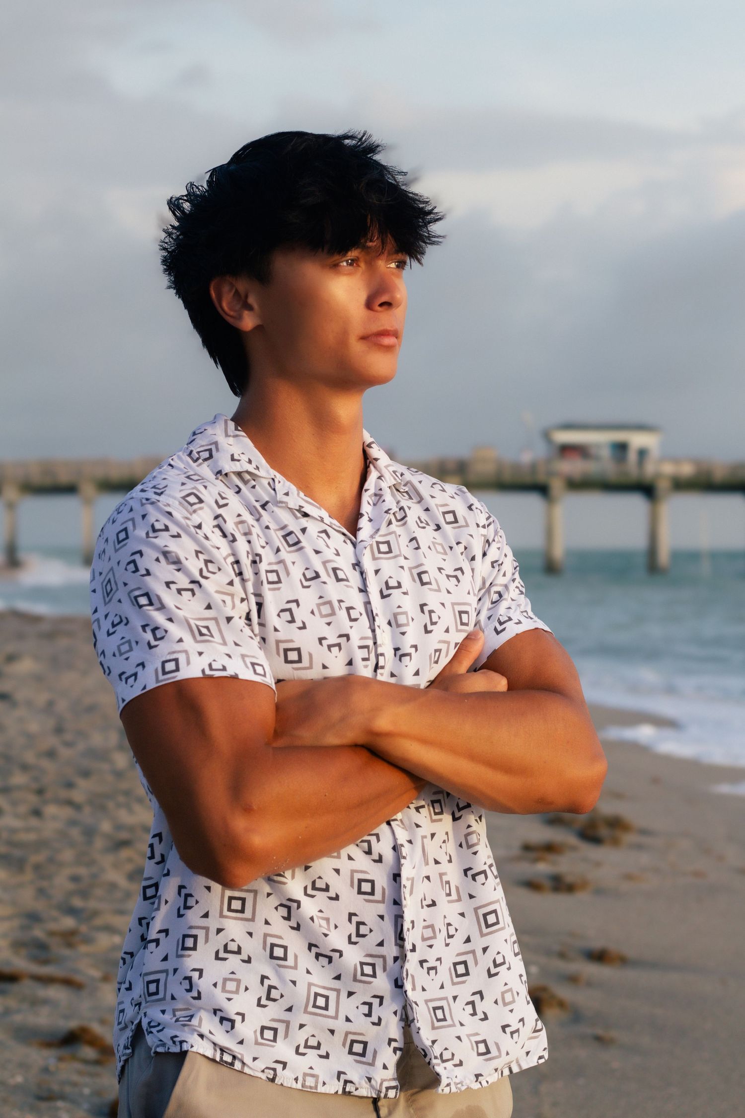 Young man in patterned shirt stands arms crossed on beach near pier at sunset.