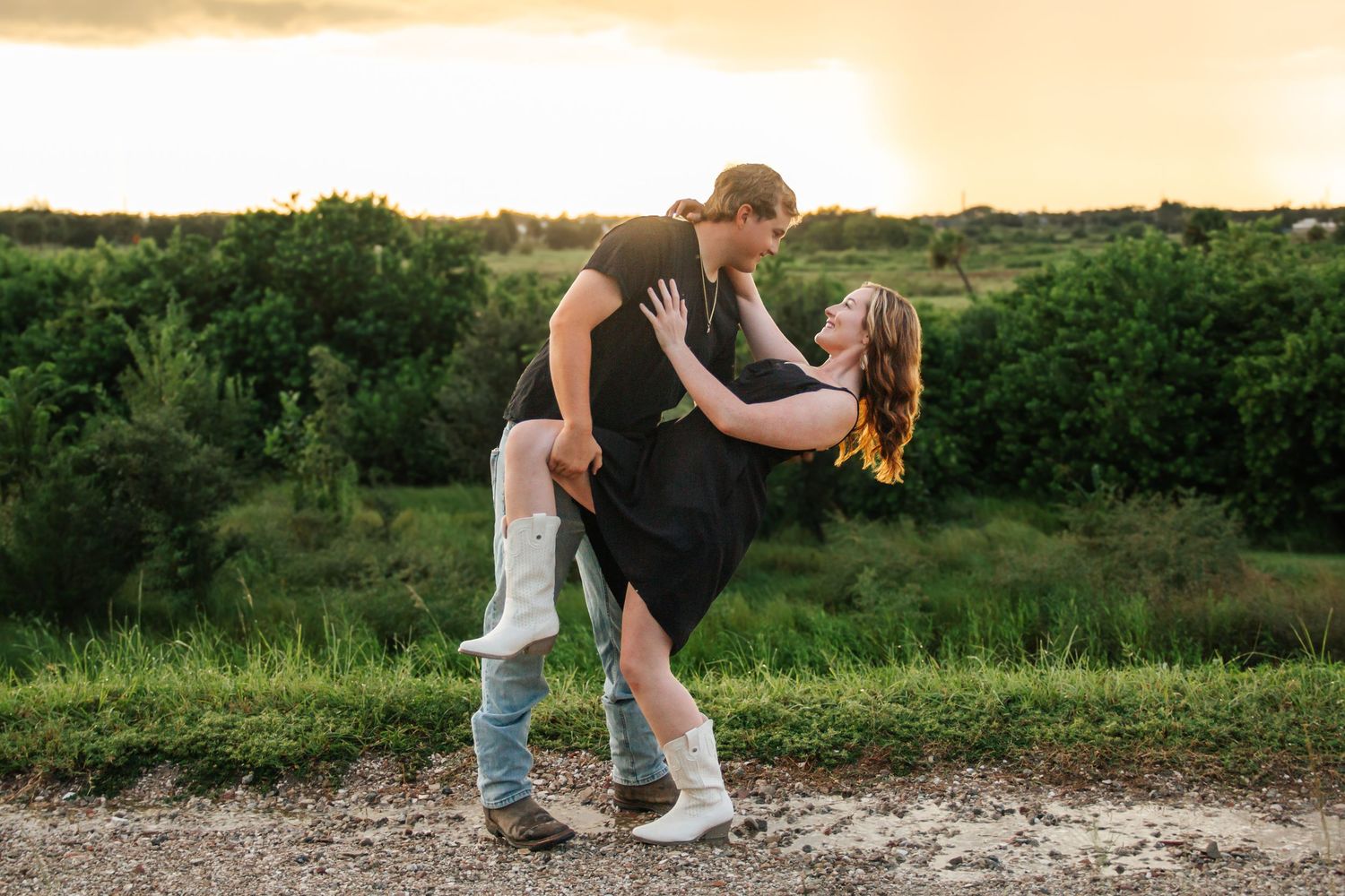 Couple dancing romantically outdoors at sunset, man dipping woman in black dress and white boots on a dirt path.