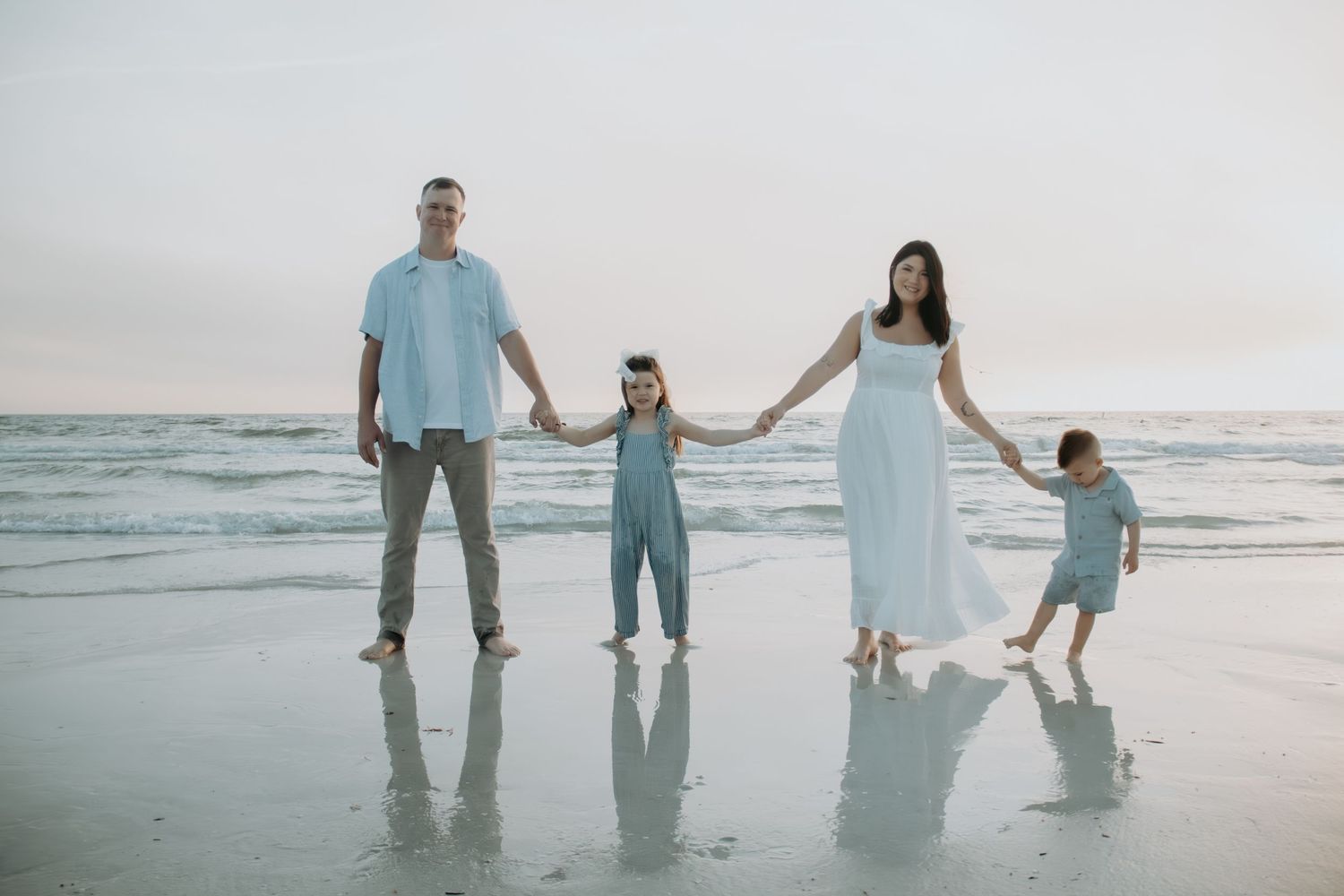Family of four holding hands on a beach at sunset, parents with two young children in light blue outfits.