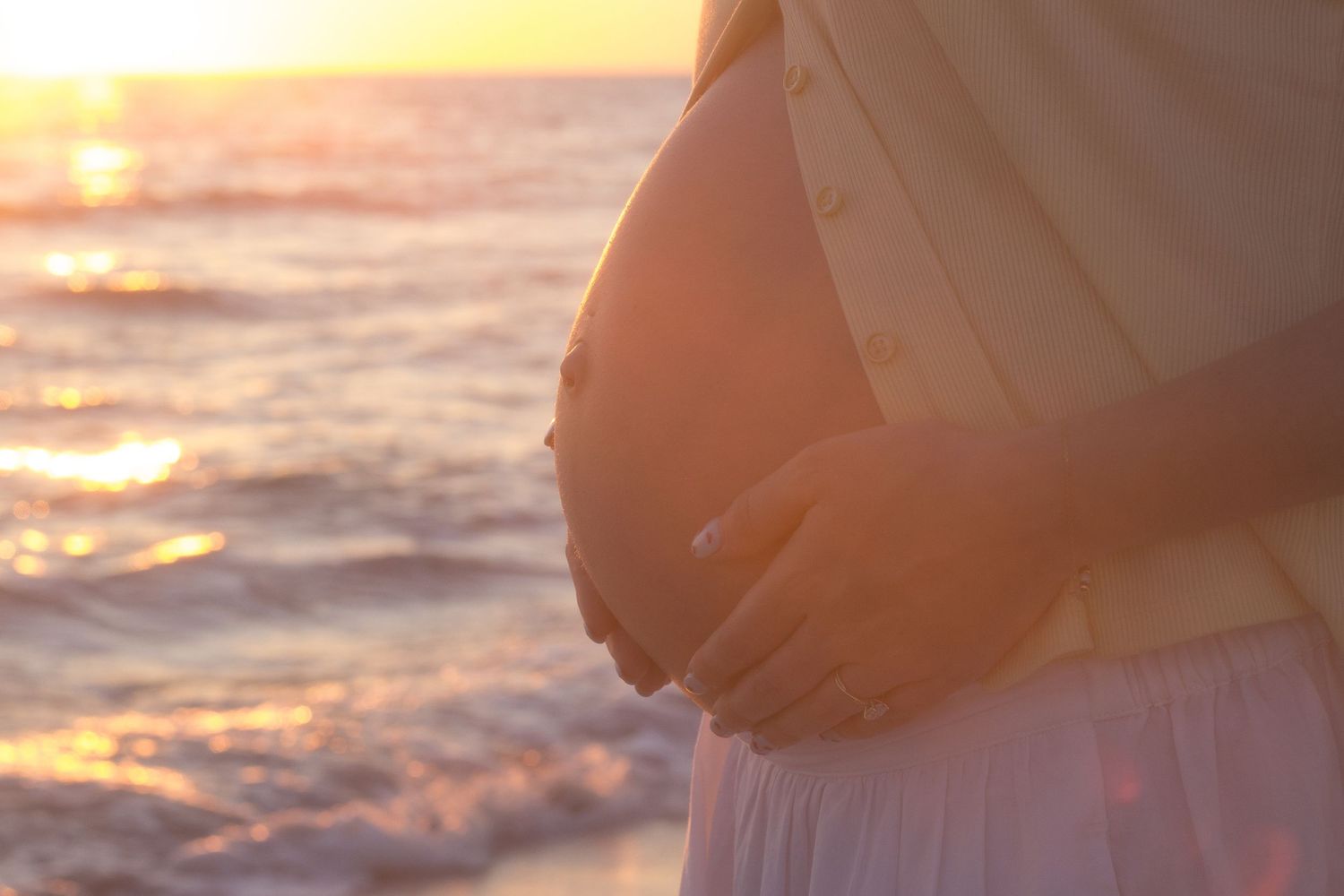 Pregnant woman cradling baby bump on beach at golden sunset with ocean waves in background.