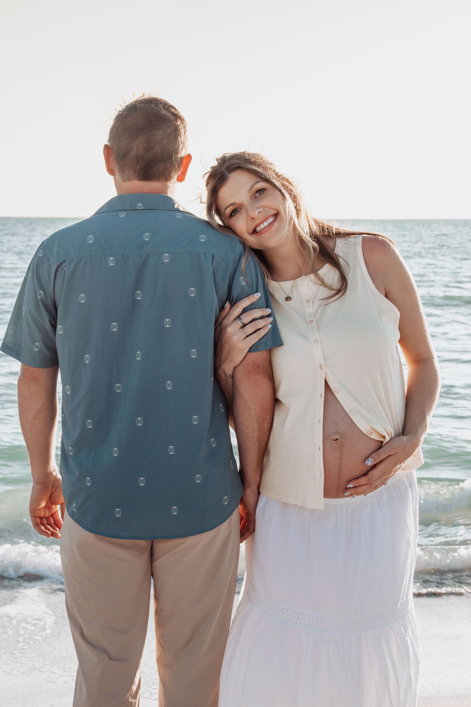 Smiling pregnant woman in white dress leans on partner's shoulder at sunset beach, cradling baby bump.
