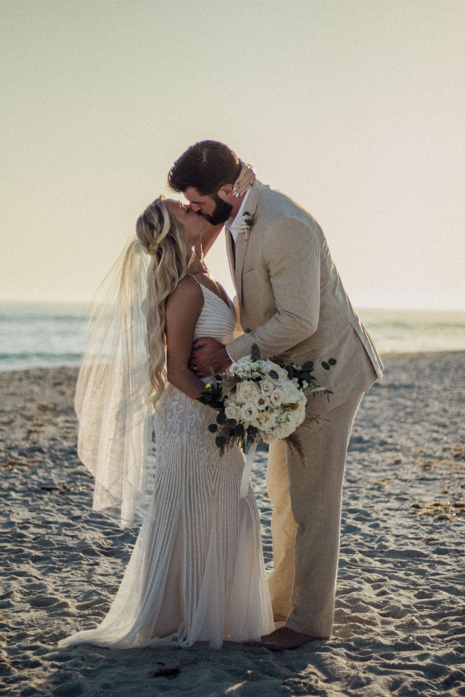 Bride and groom sharing a romantic kiss on the beach at sunset, bride in white gown with veil holding bouquet.