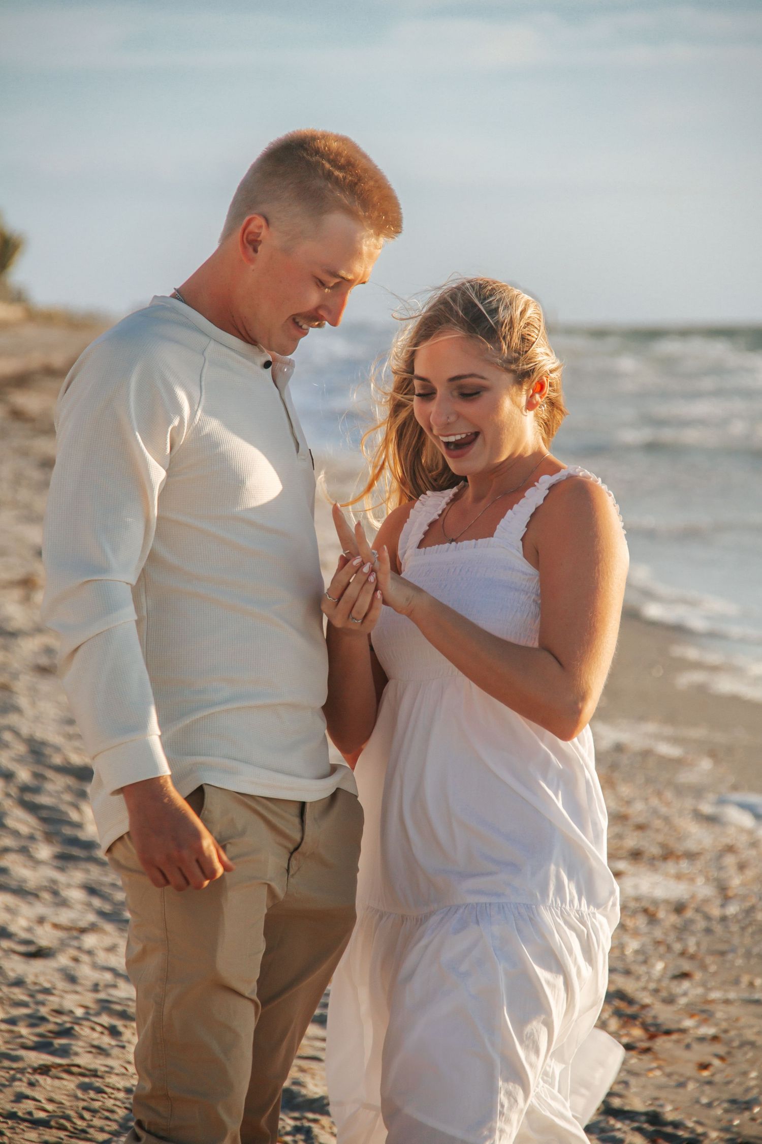 Happy couple admiring engagement ring on beach at sunset, woman in white dress expressing joy.