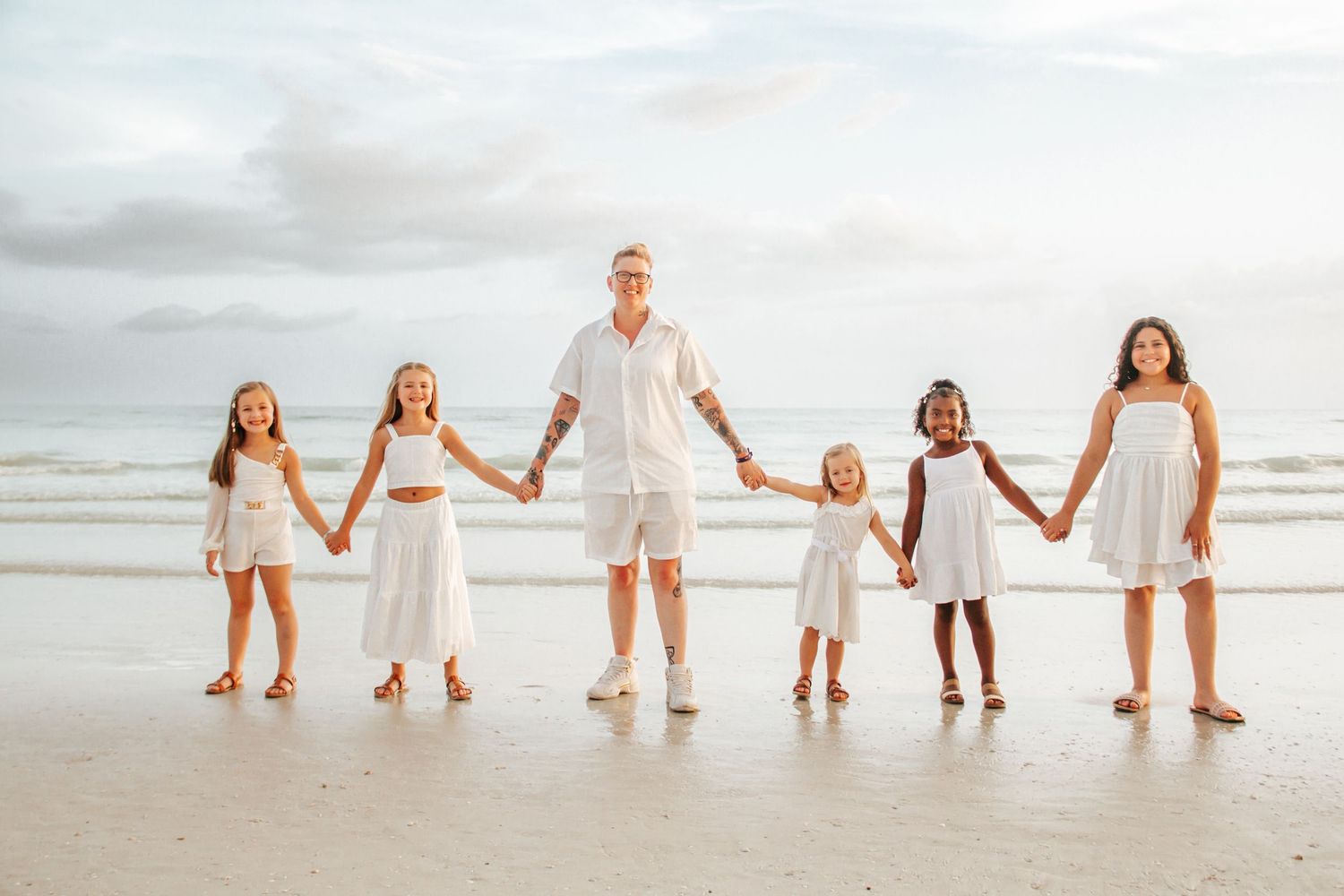 Family of six holding hands on a beach, dressed in white outfits, smiling with ocean waves in the background.