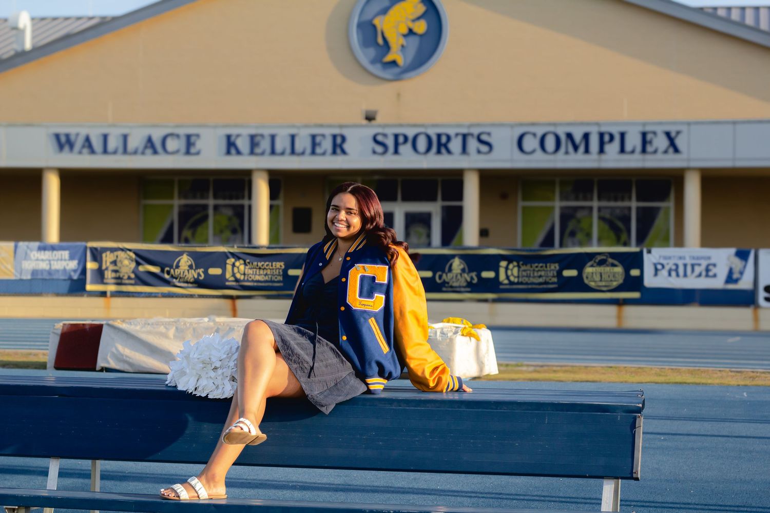 Young woman in varsity jacket sitting on bleachers at Wallace Keller Sports Complex during golden hour.