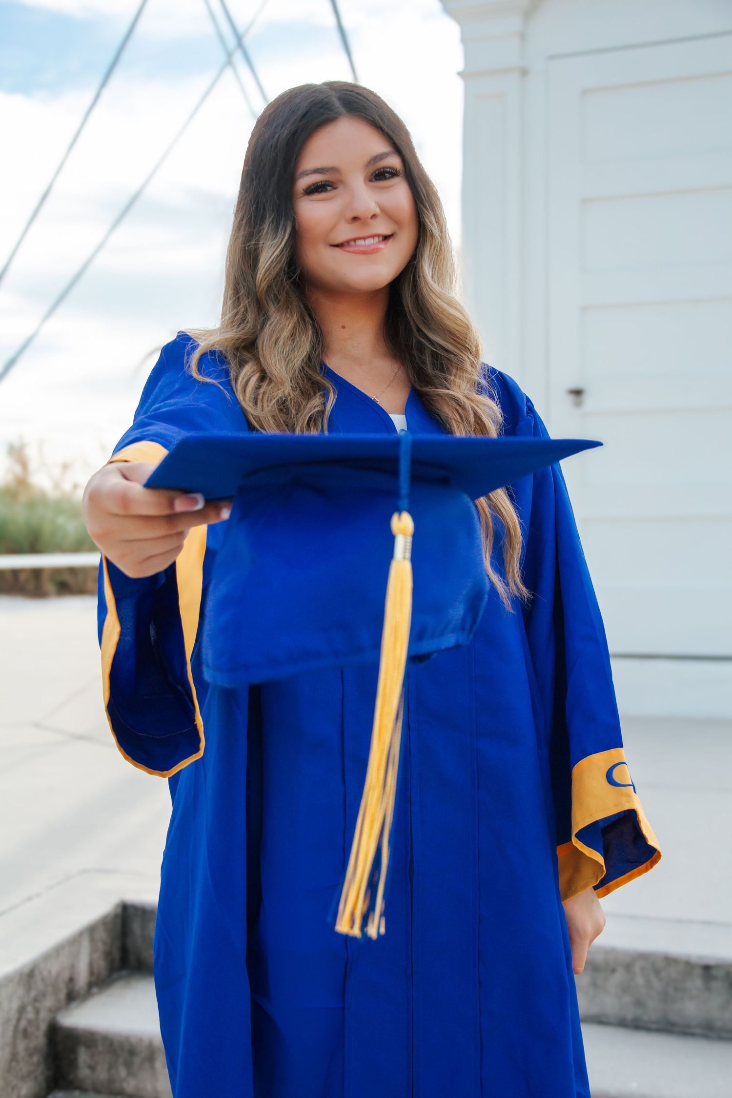 Smiling young woman in blue and gold graduation cap and gown holding mortarboard toward camera outdoors.