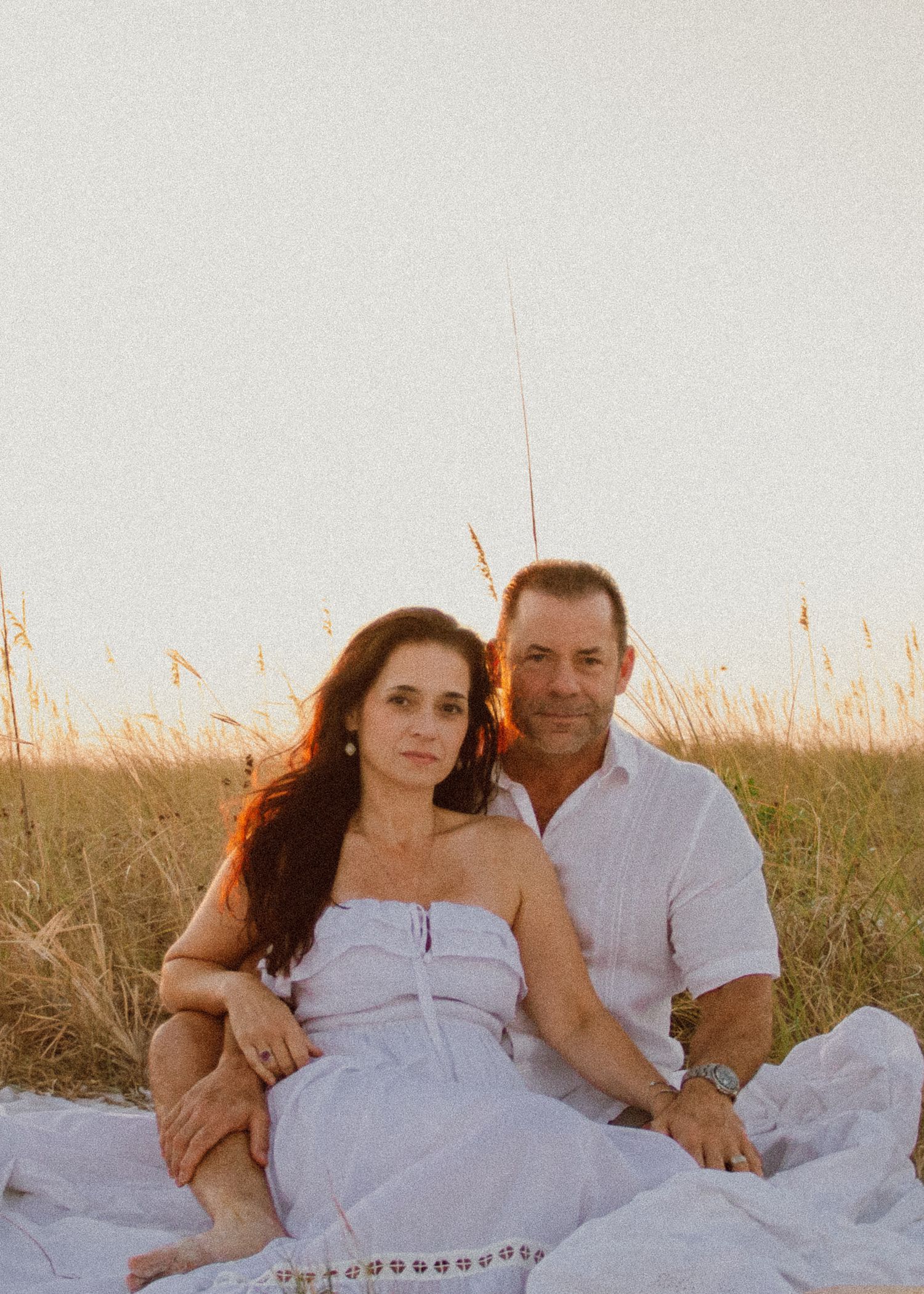 Couple sitting together on a white blanket in golden beach dunes at sunset, woman in white dress, man in linen shirt.