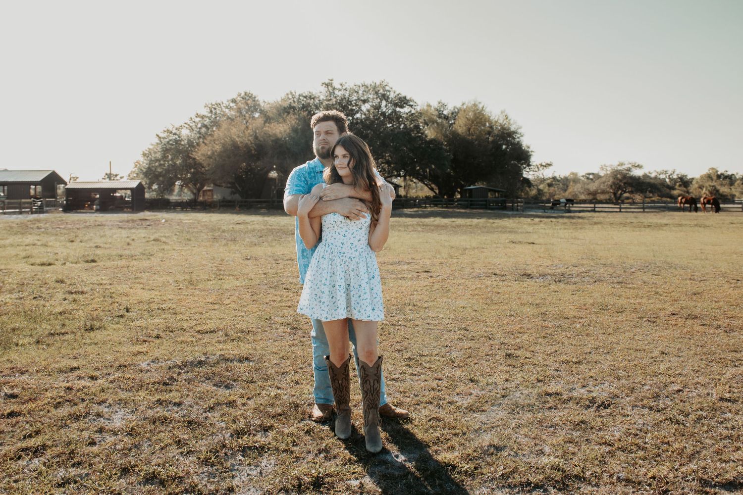 Couple embracing in a sunny open field, woman in floral dress and cowboy boots, man in blue shirt, rustic farm background.